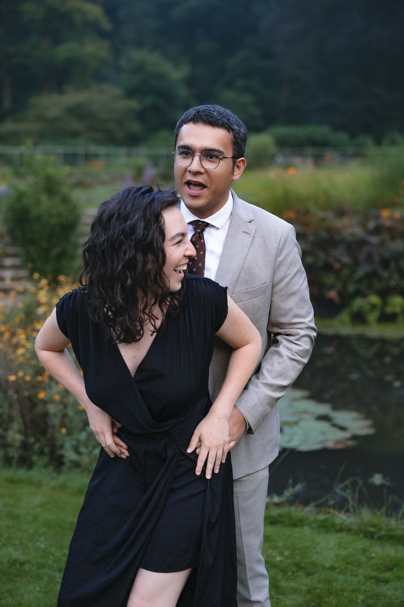 A couple embraces and laughs together in a garden setting, with the woman wearing black and the man in a light gray suit.