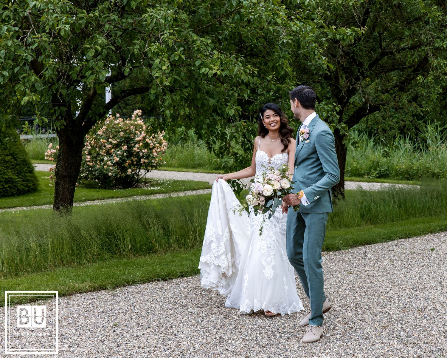 A couple strolls along a gravel path surrounded by lush gardens and greenery.

- BU Photography -