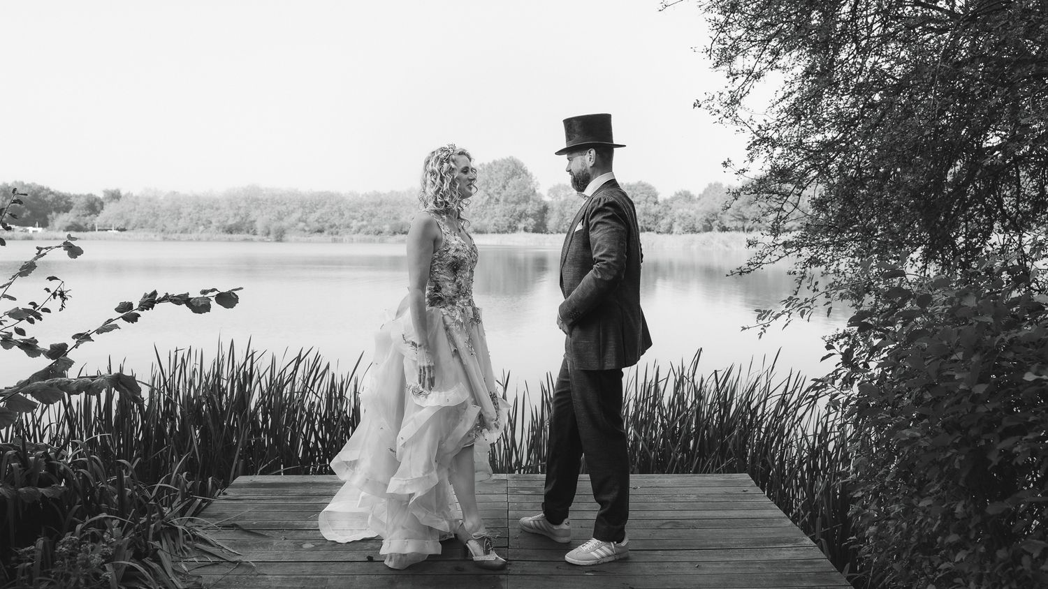 A black and white wedding photo showing a couple standing on a wooden dock by a serene lake surrounded by nature.