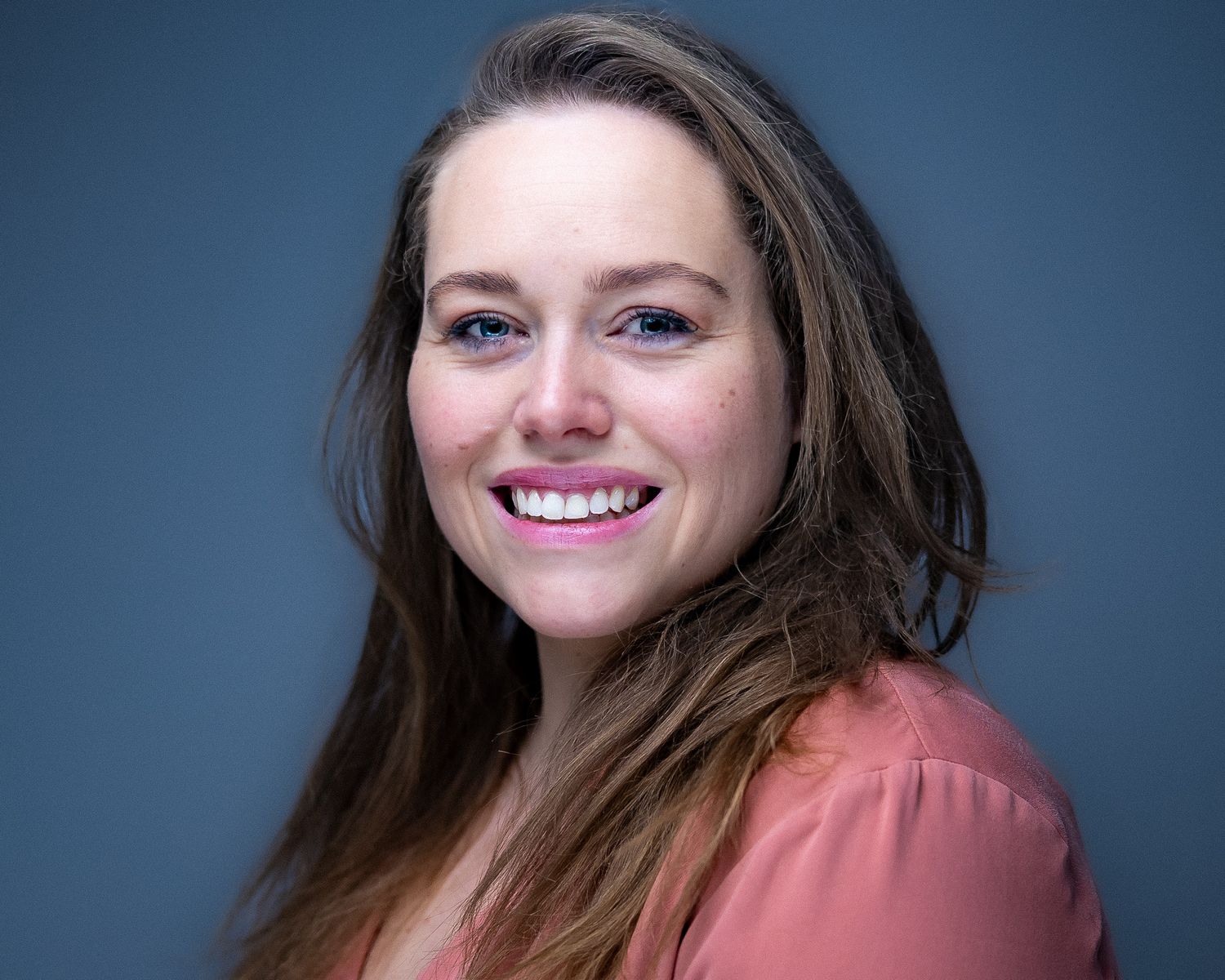 Professional headshot portrait against blue background showing warm smile and wearing a coral-colored blouse.