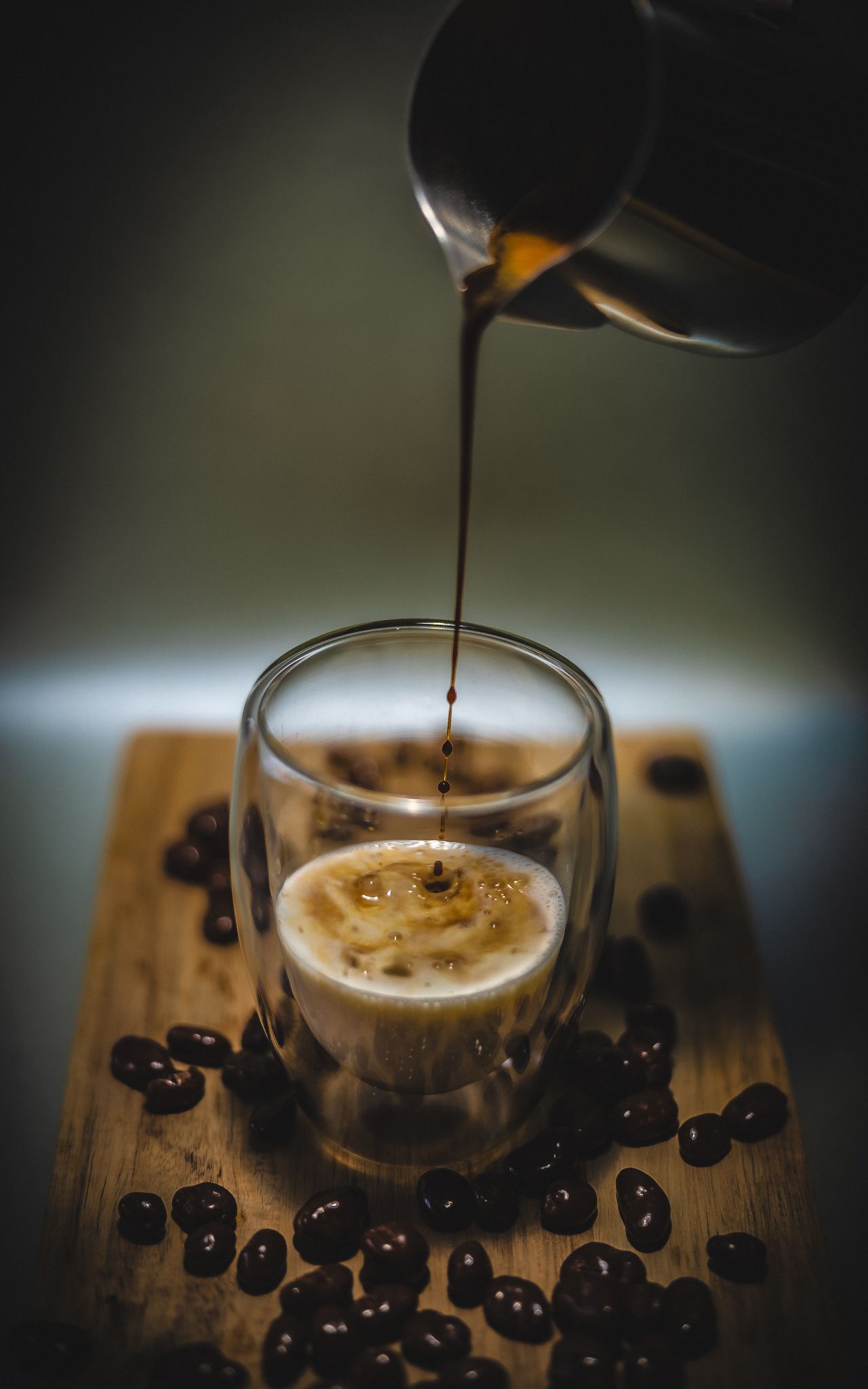 Fresh espresso being poured into a glass cup surrounded by scattered coffee beans on a rustic wooden board.