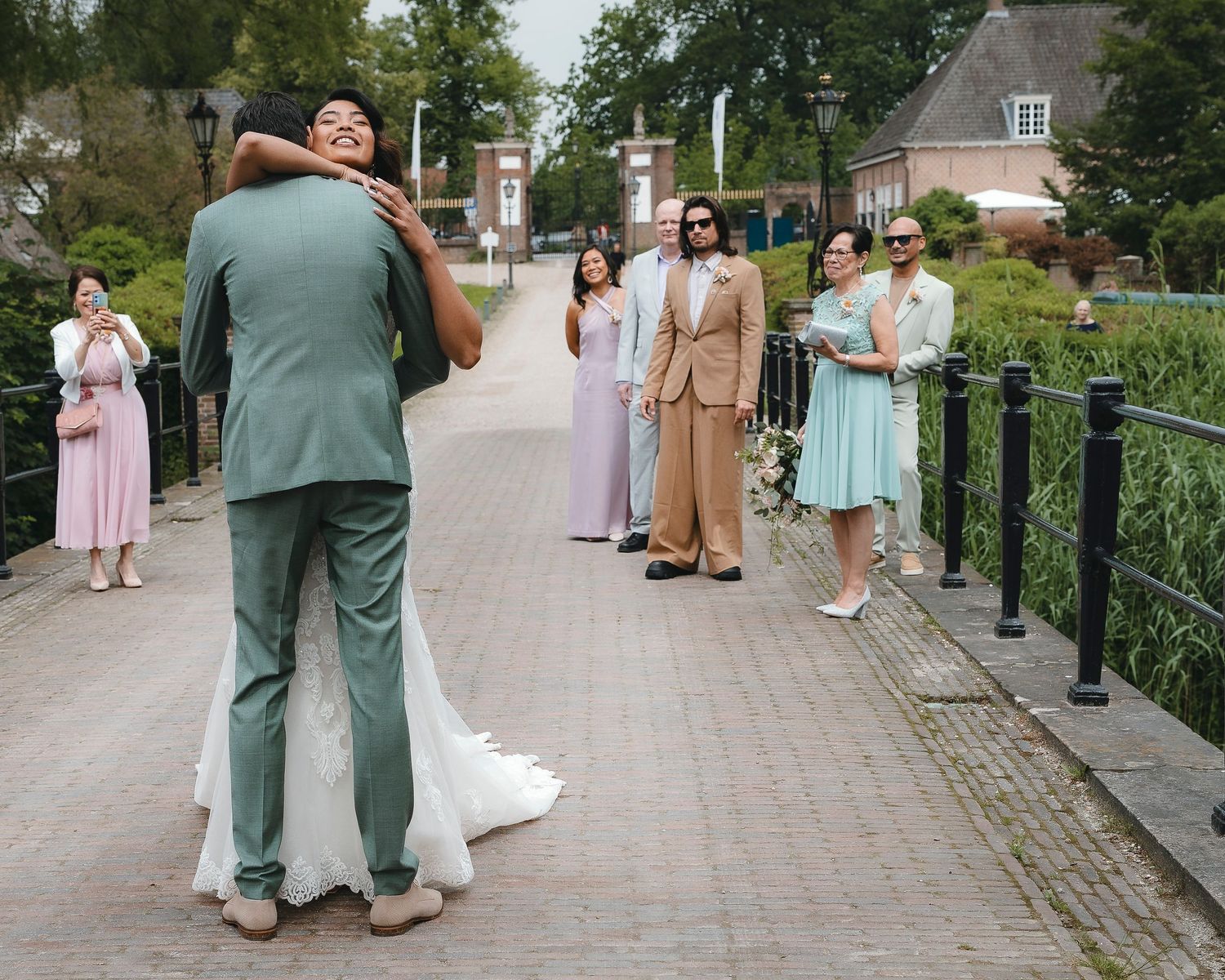 A wedding party stands on a brick paved path as guests wait for photos to be taken in an outdoor garden setting.