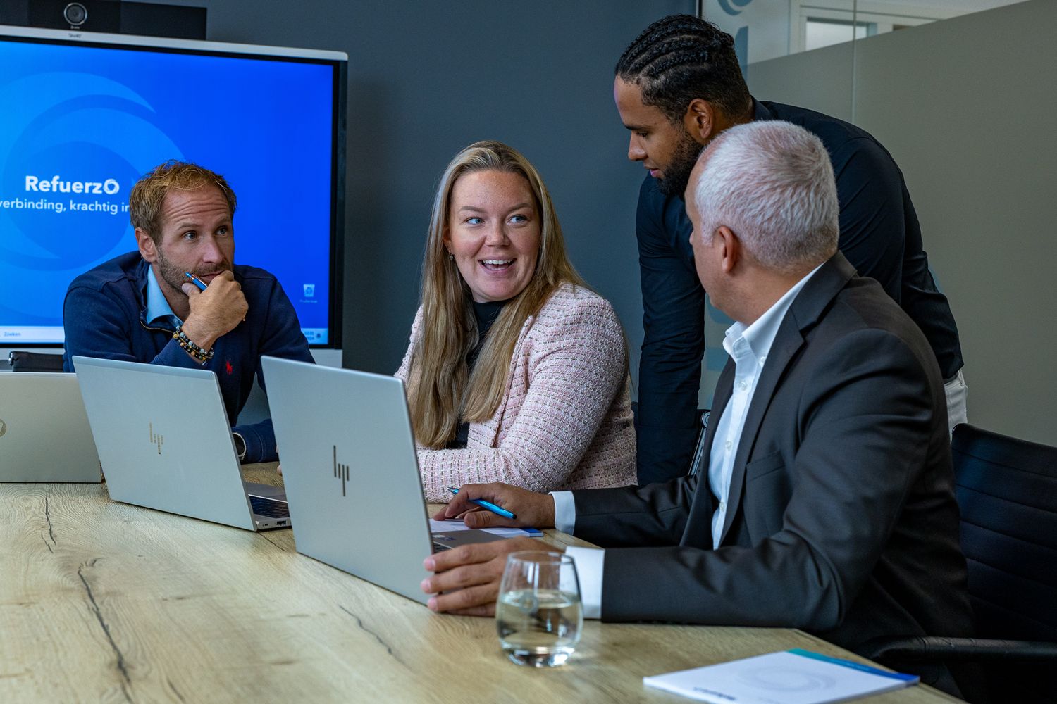 Business team collaborates at a modern conference table while reviewing data on a laptop near a large blue display screen.