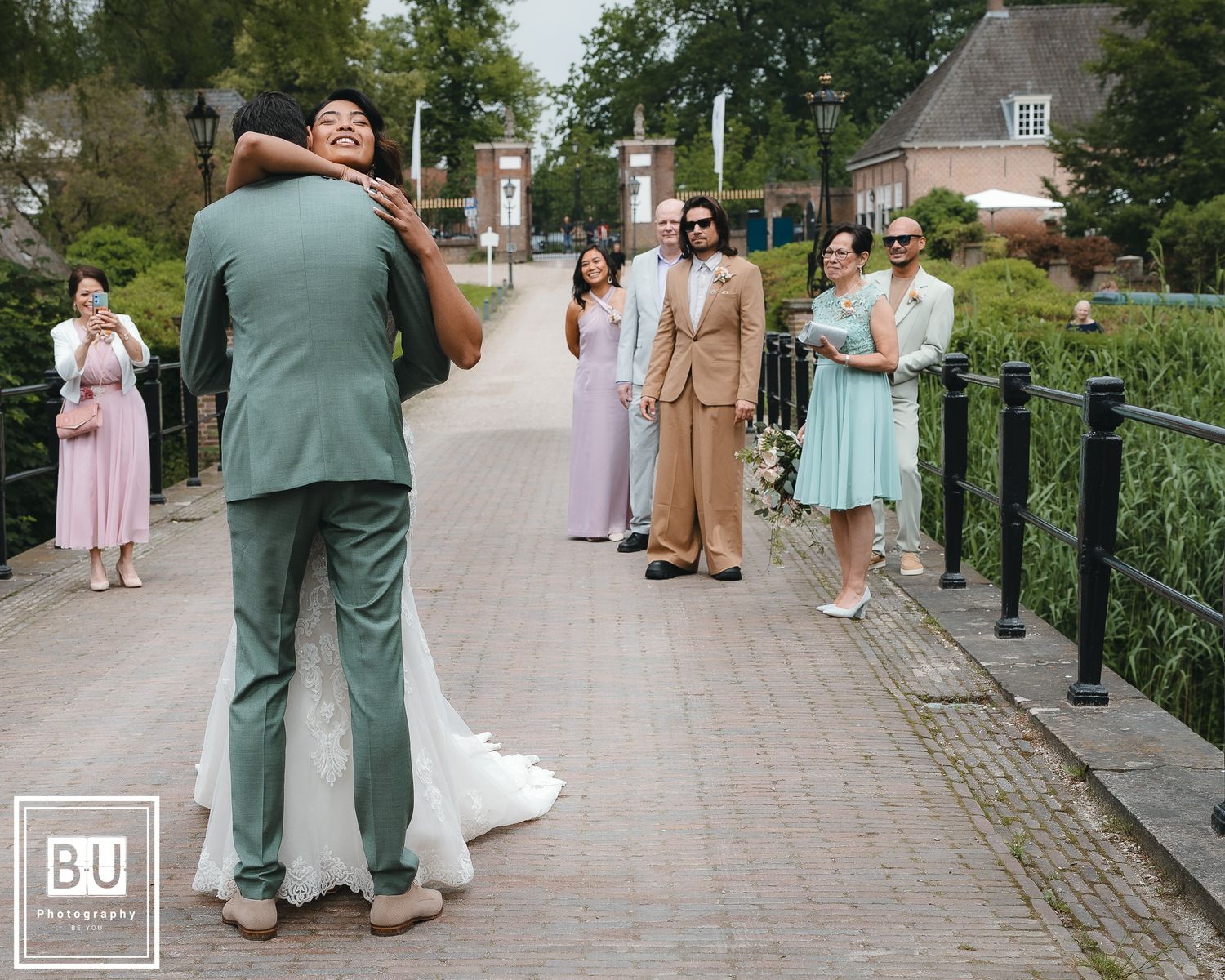 Wedding party gathers on a brick pathway lined with greenery while couple embrace in foreground.