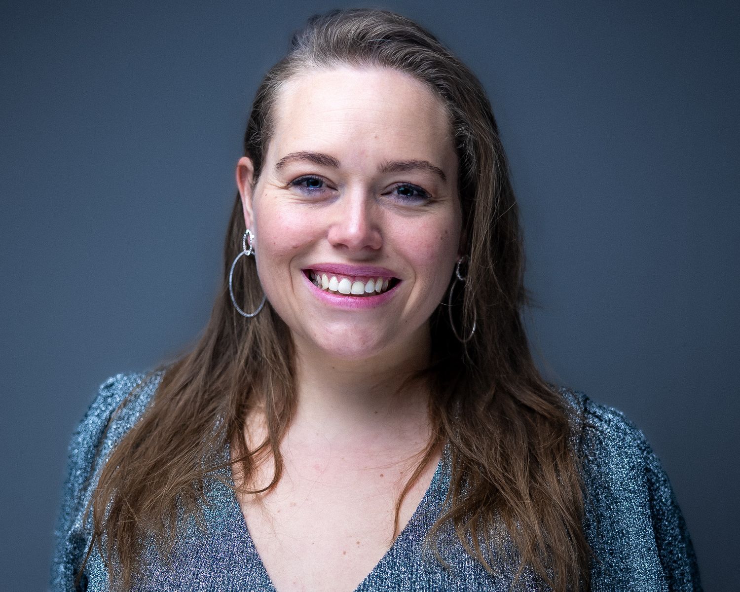Professional headshot of someone with brown hair wearing a blue top and hoop earrings with a warm smile against dark background.