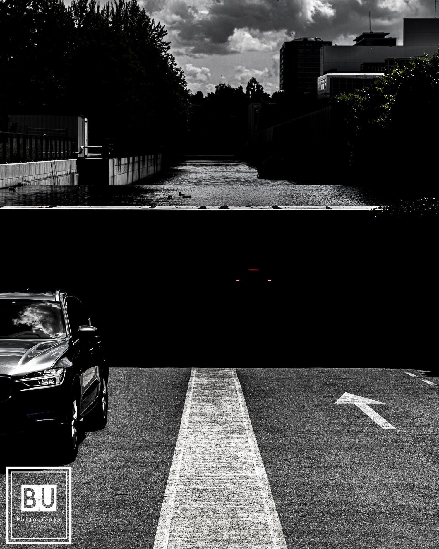 Black and white urban parking lot with a car and directional arrow under dramatic cloudy skies.