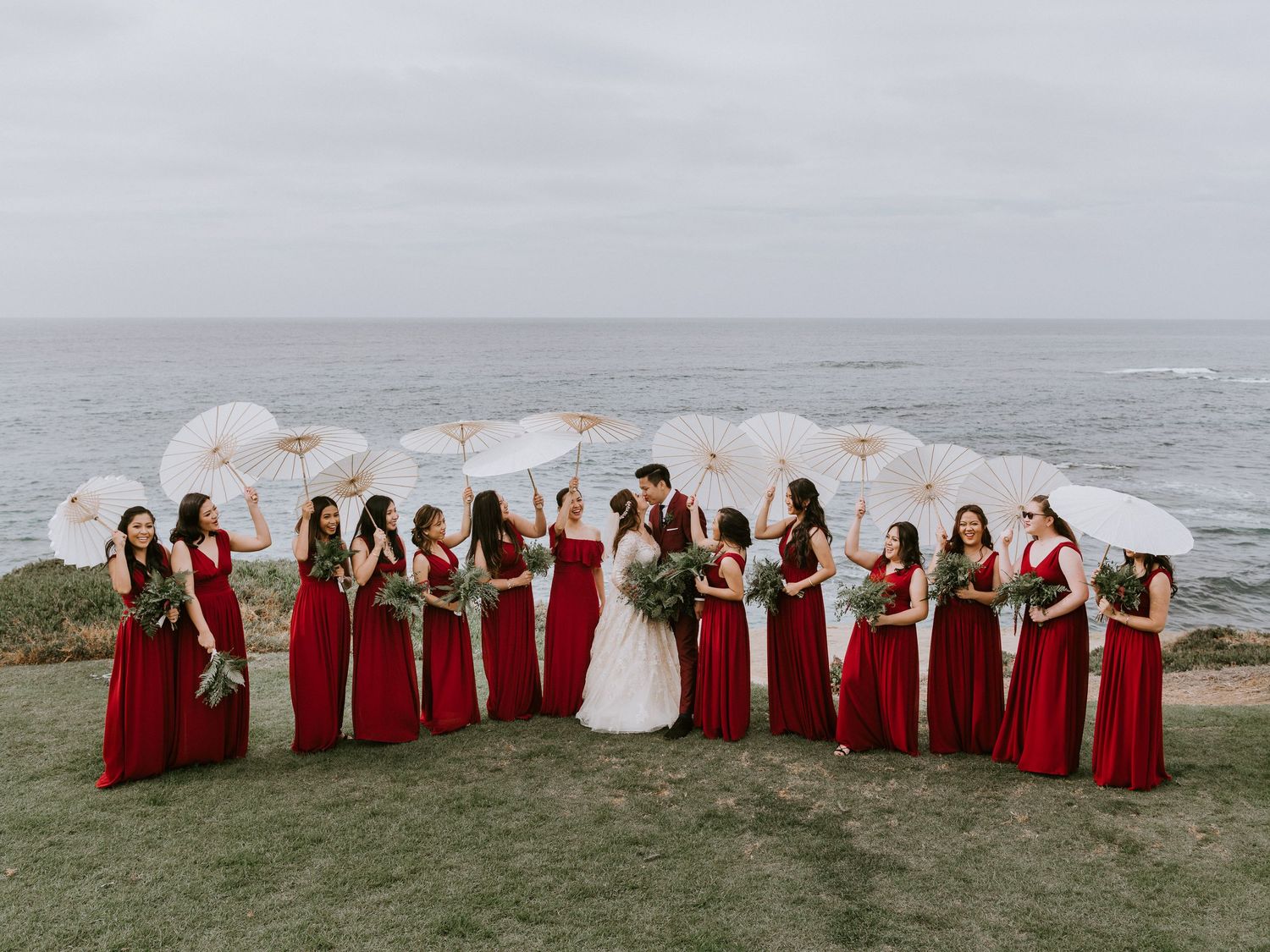 Wedding party poses with white parasols and red bridesmaid gowns at stunning oceanside venue.