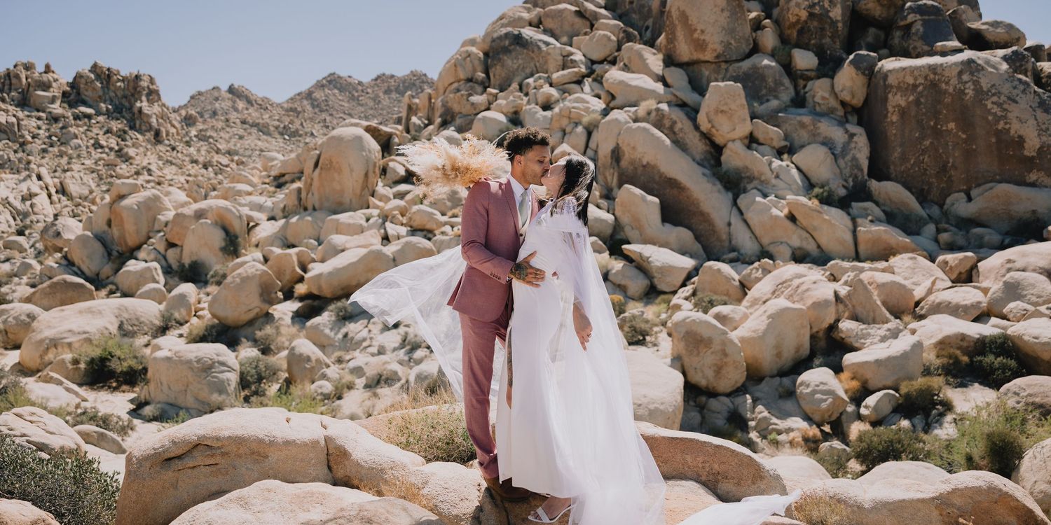 Couple sharing a romantic kiss in a desert landscape, bride in white gown and groom in pink suit among large boulders.