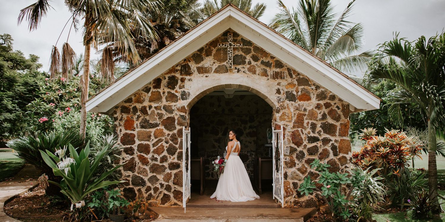 Bride in white gown standing in archway of rustic stone chapel surrounded by tropical greenery.