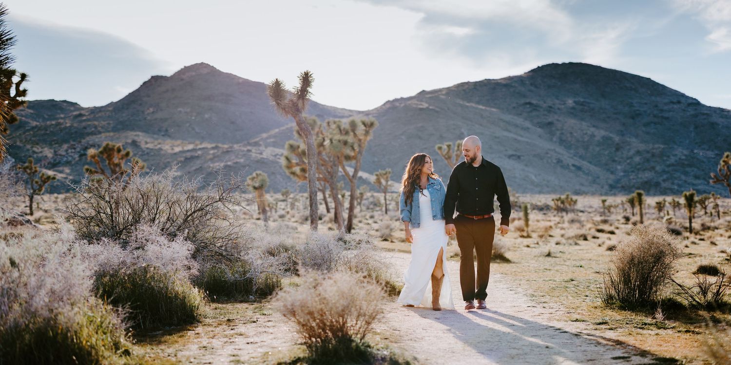 Couple walks hand in hand on a desert path surrounded by Joshua trees and mountains in golden light.