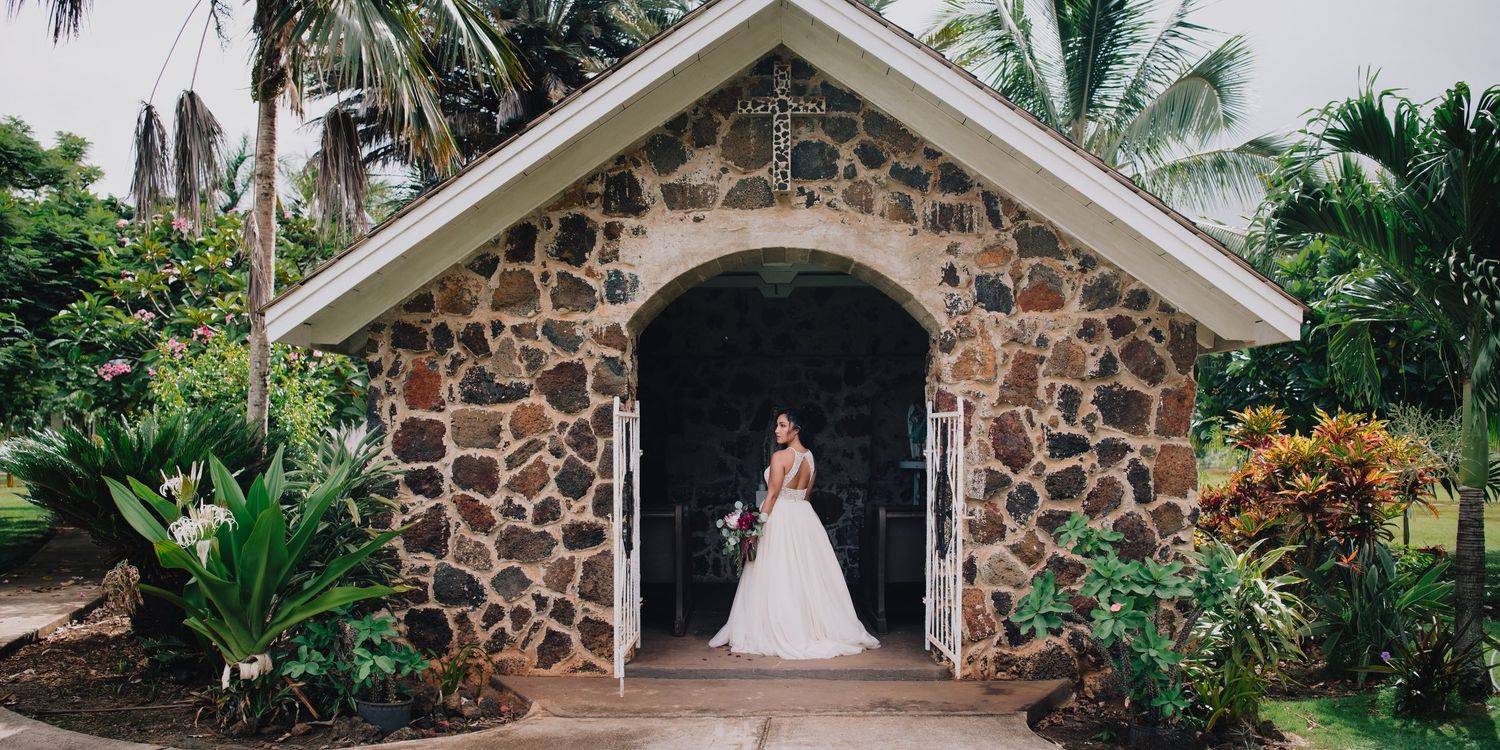 Bride in white wedding gown standing in archway of rustic stone chapel surrounded by tropical greenery.