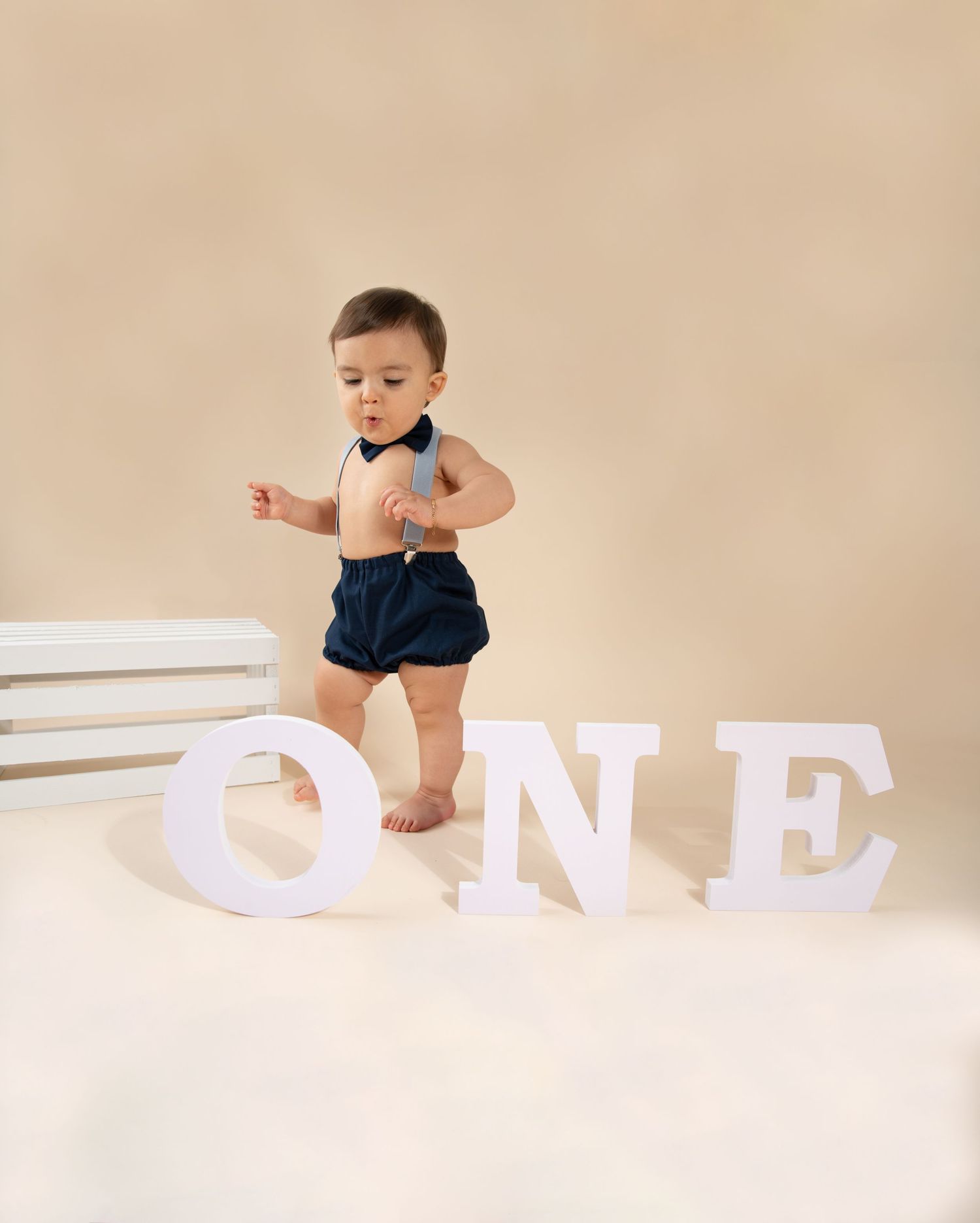 A toddler in a navy blue bow tie and shorts standing next to white letters spelling 'ONE' against a beige backdrop.
