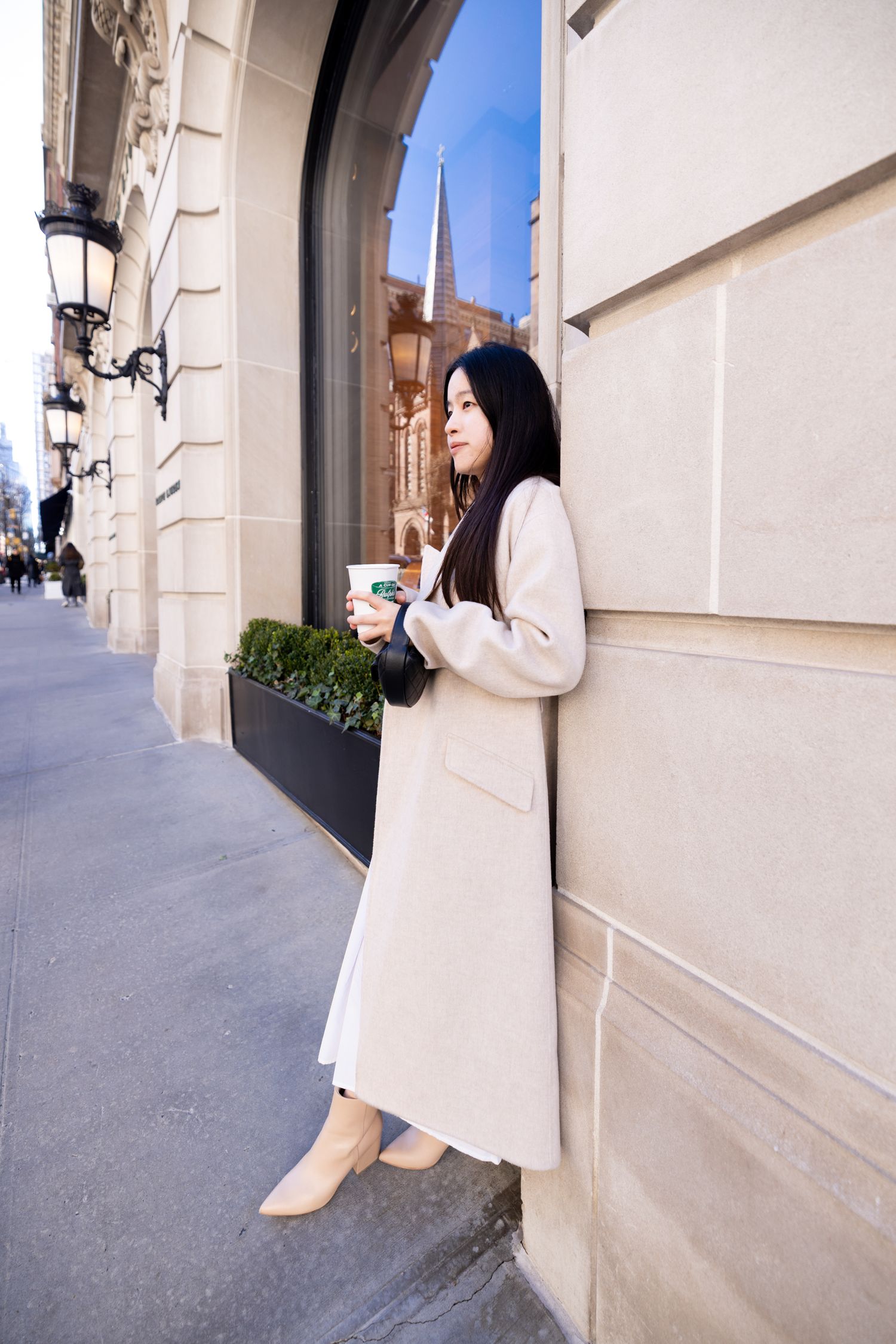 Person in cream colored long coat leans against building exterior holding coffee cup on urban street with architecture.