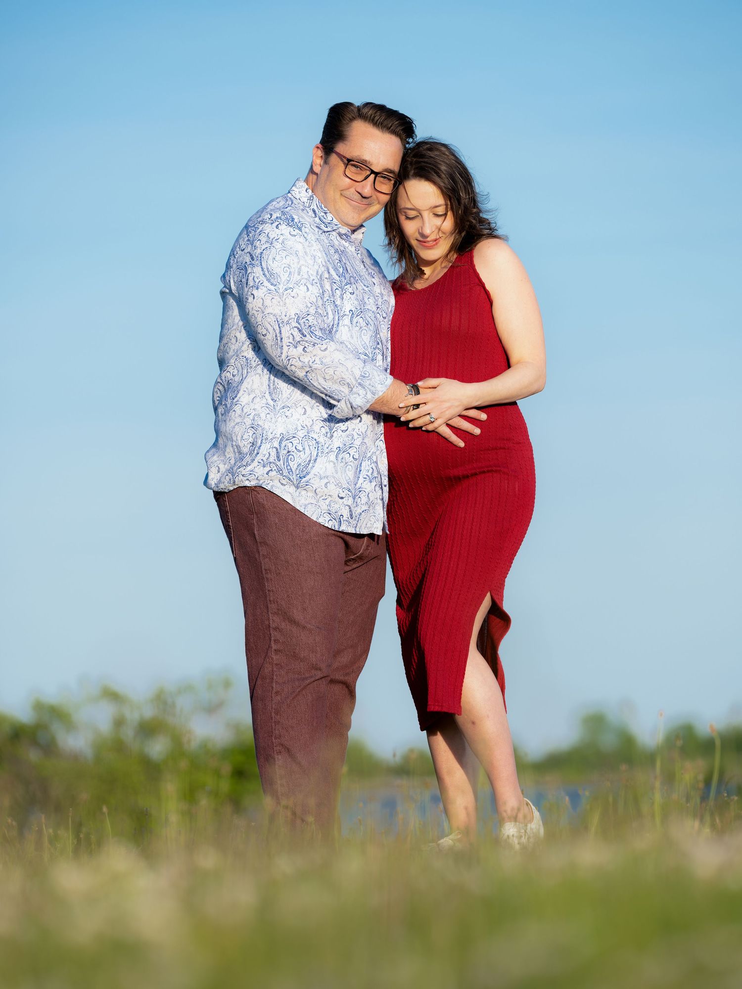 A couple embraces outdoors in coordinating outfits, with one wearing a patterned shirt and the other in a red dress.