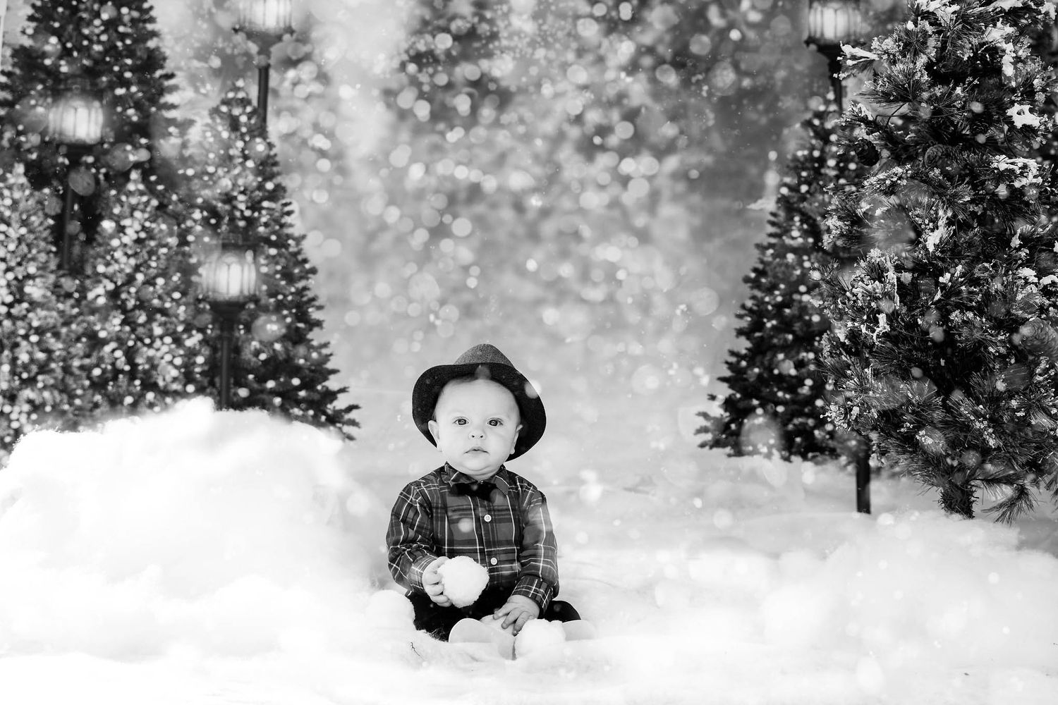 A small child sits in the snow surrounded by evergreen trees in a dreamy black and white winter scene.