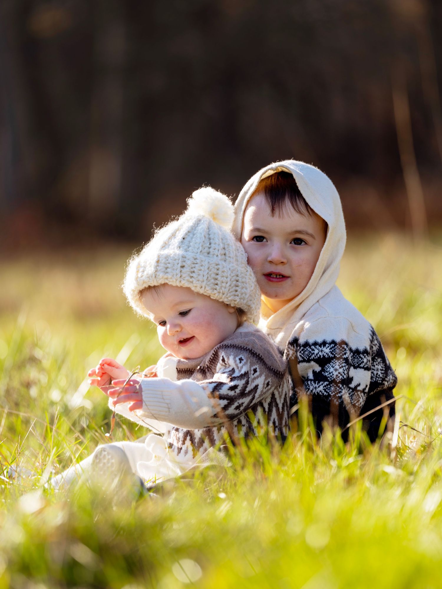 Two young children sit together in sunlit grass on a crisp autumn day wearing cozy knit winter clothing.