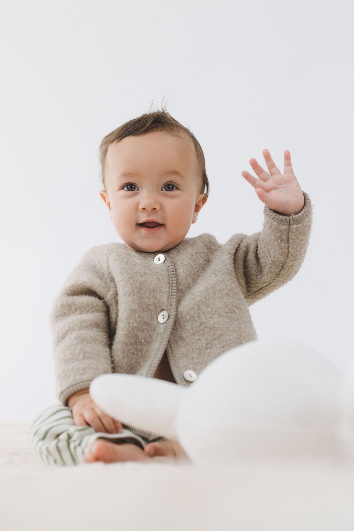Sweet baby in gray cardigan sweater waves and smiles while lying on white bedding in a series of photos.