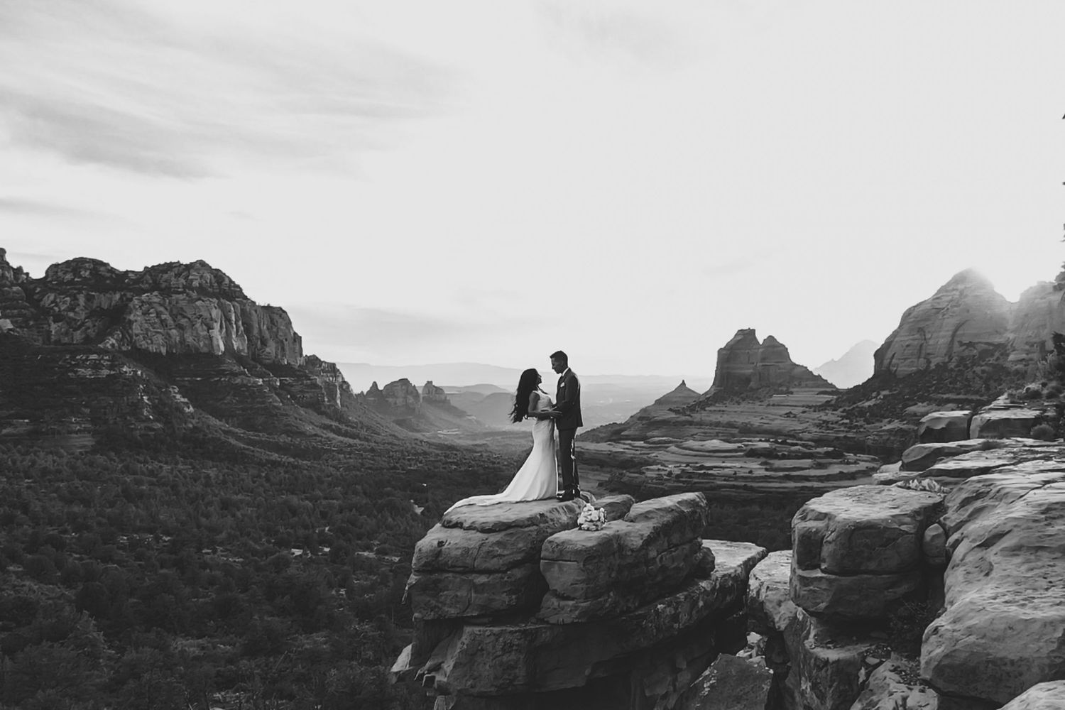 Black and white photo of a couple in wedding attire standing on a rocky cliff overlooking Sedona, Arizona red rock landscape.