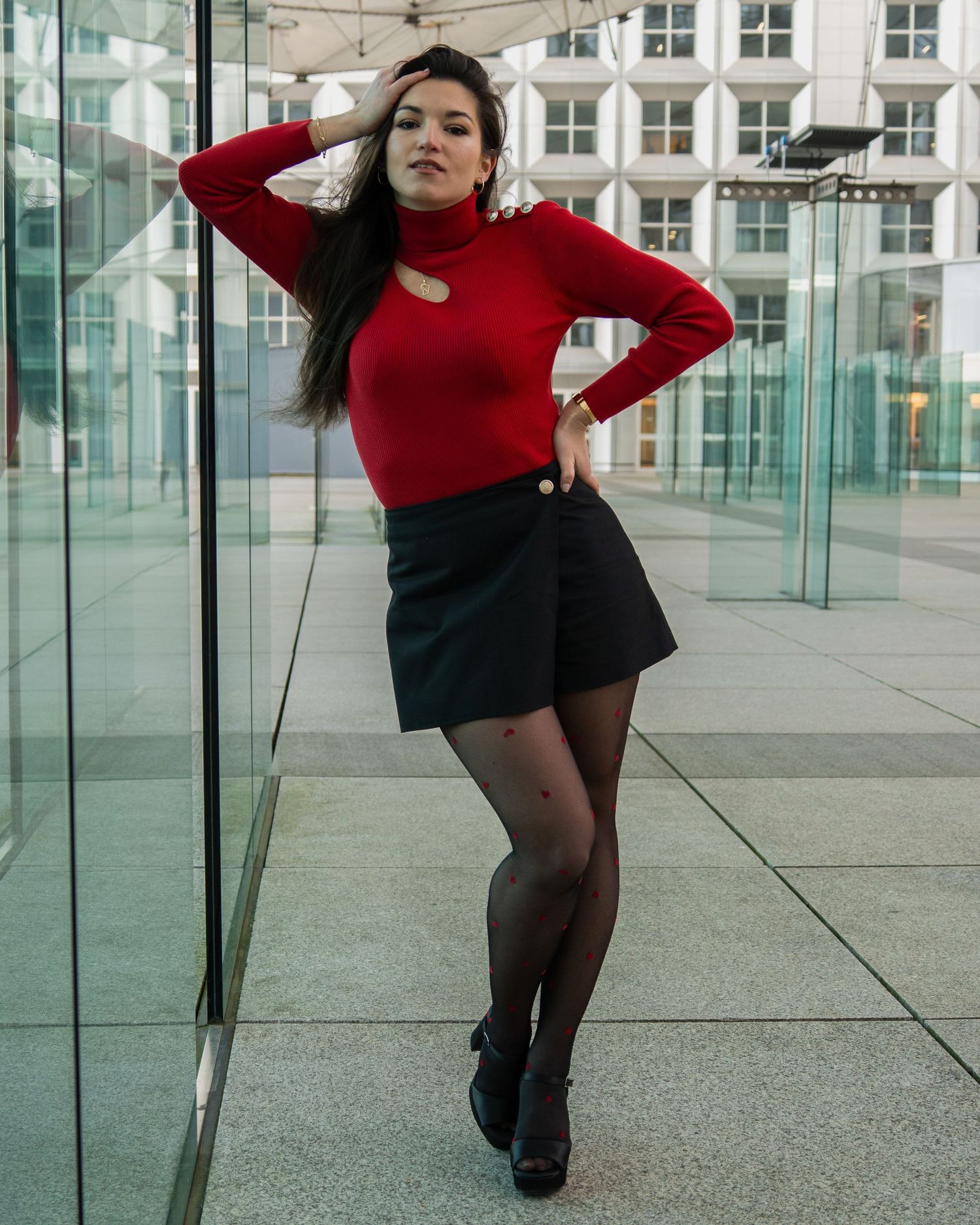 Portrait de mode contre un bâtiment en verre avec un pull à col roulé rouge et un ensemble mini-jupe noire sur Paris.