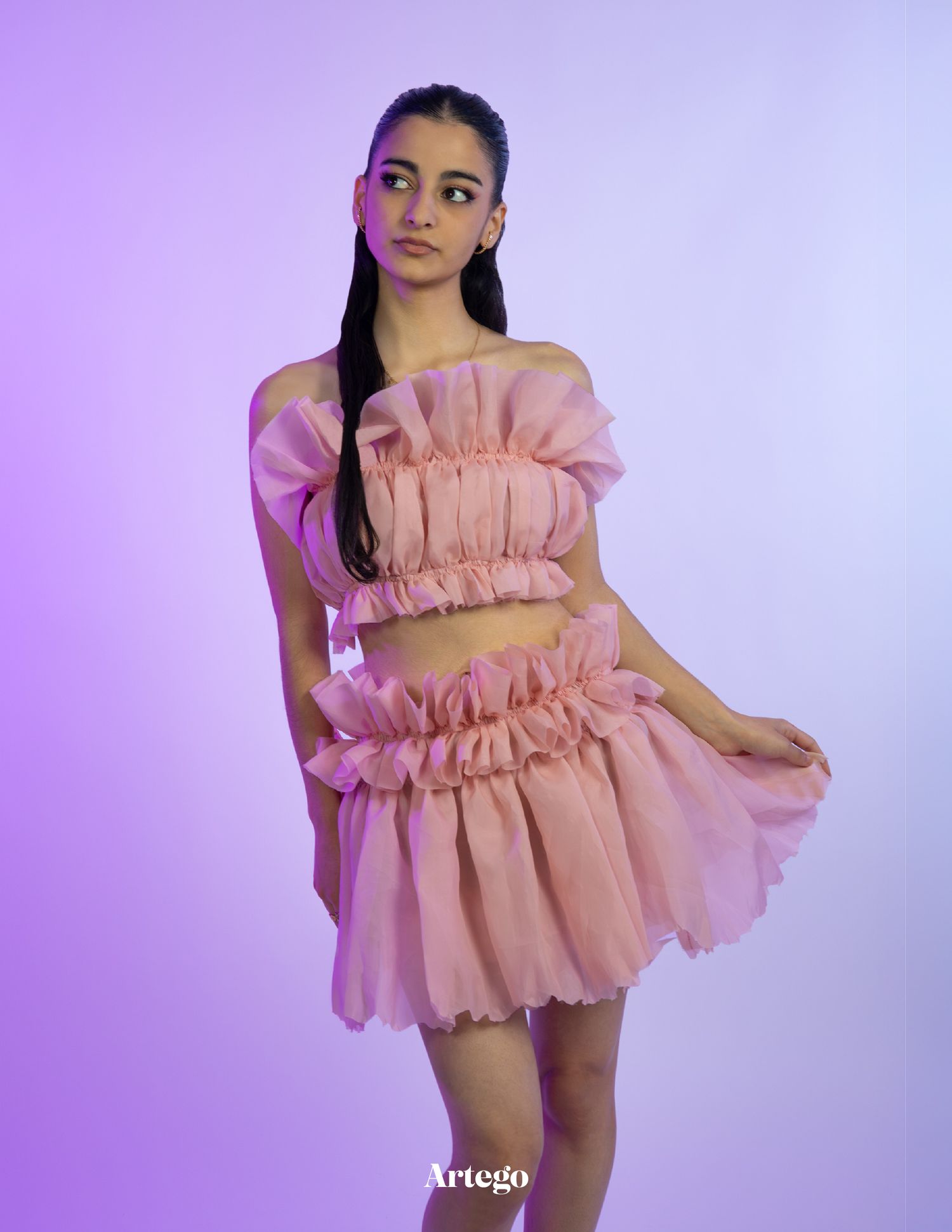 Model wearing pink ruffled two-piece dress poses against purple-lit studio background.