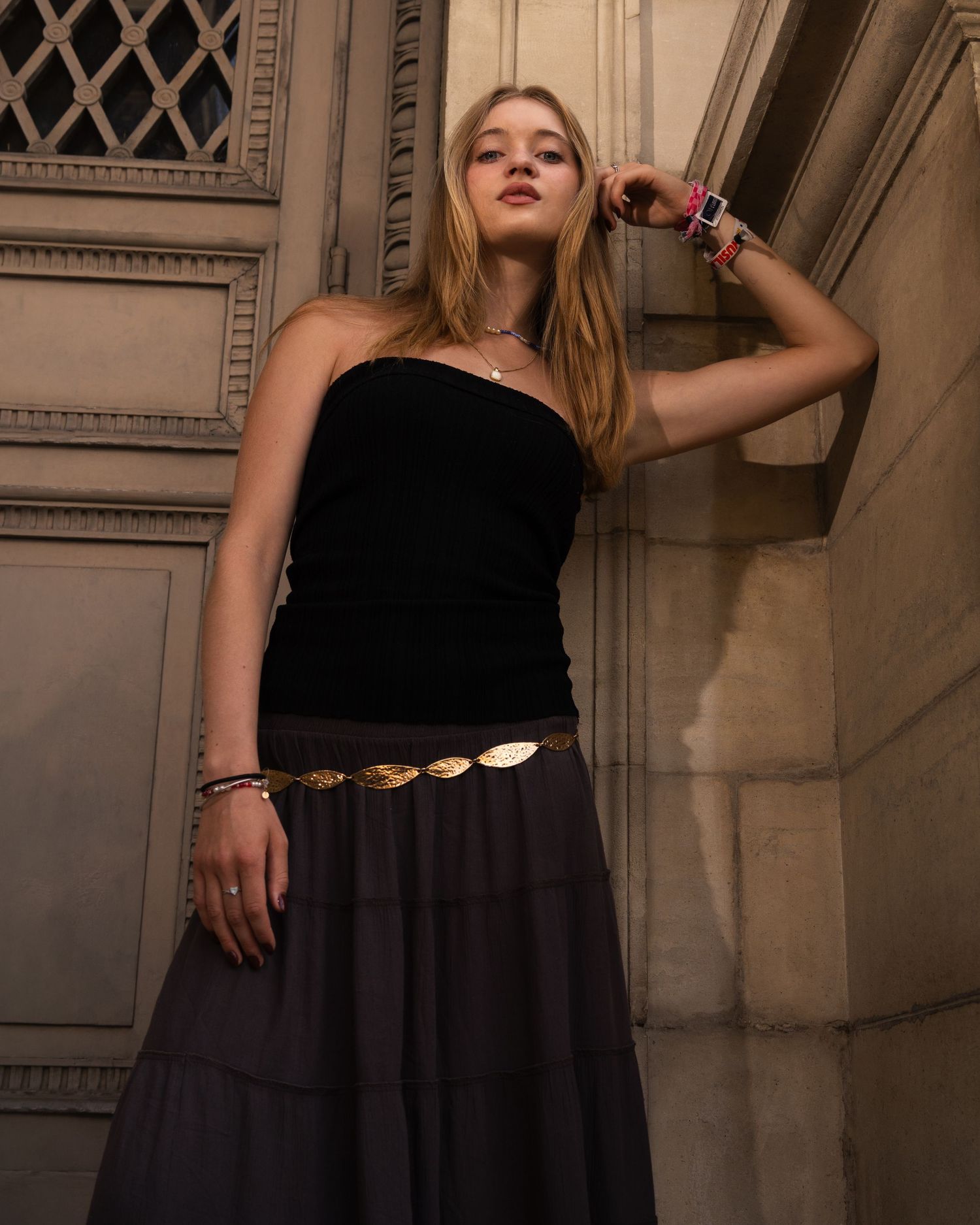 Young woman in black strapless top, tiered maxi skirt, and gold coin belt poses against ornate stone wall.
