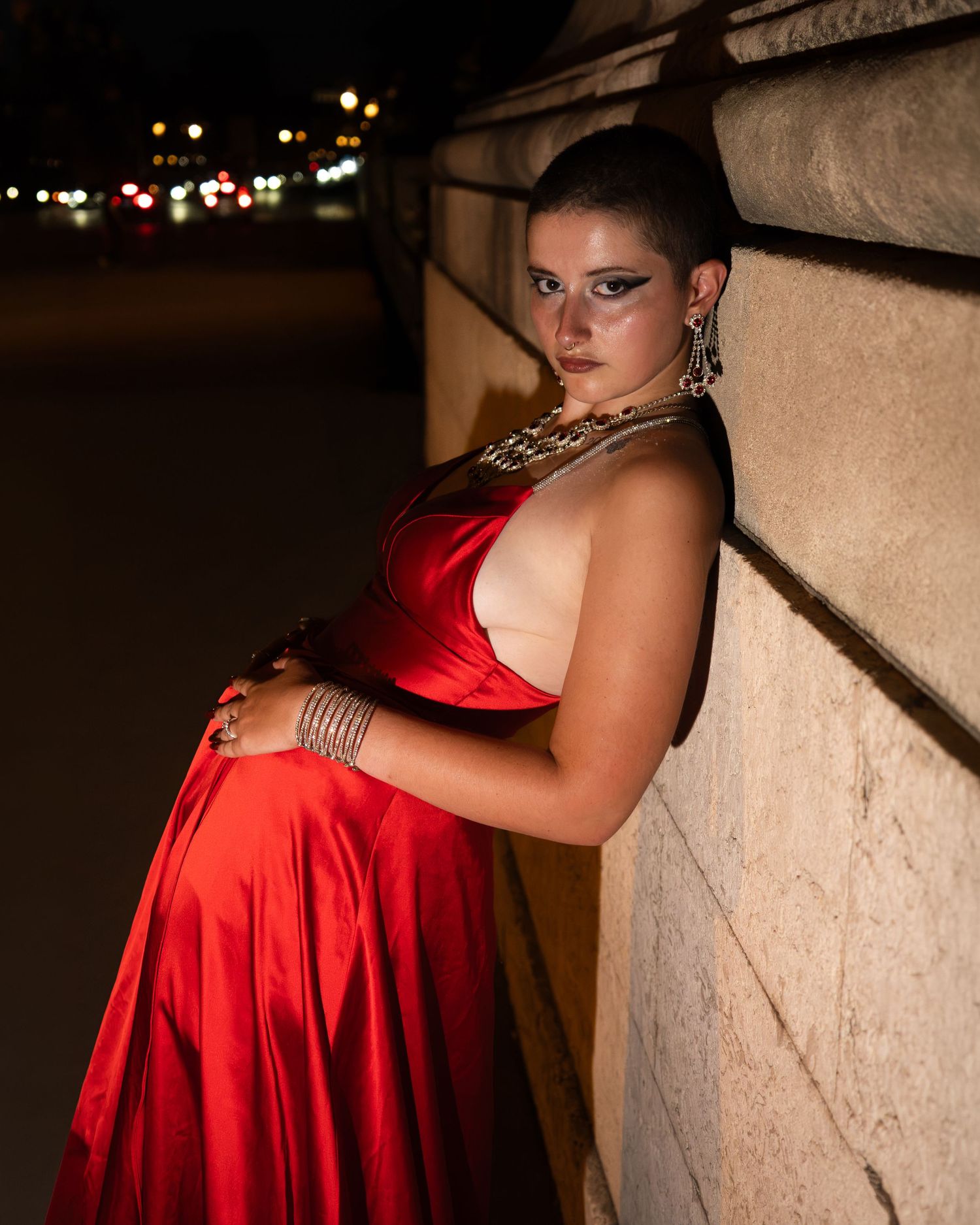 Woman in red satin gown with gold jewelry poses against stone column at night.