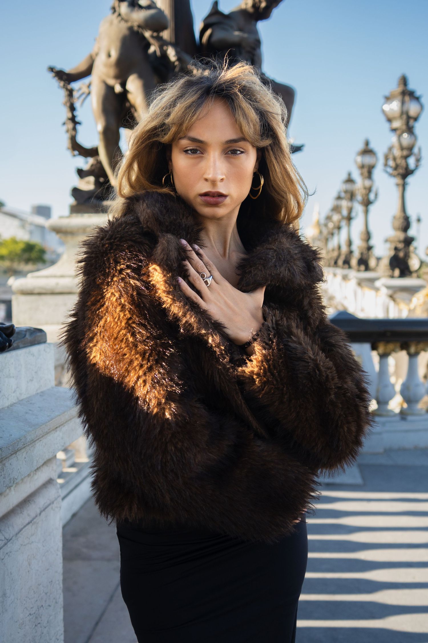 Woman in brown fur coat posing on elegant Parisian bridge with ornate lampposts and sculptures.