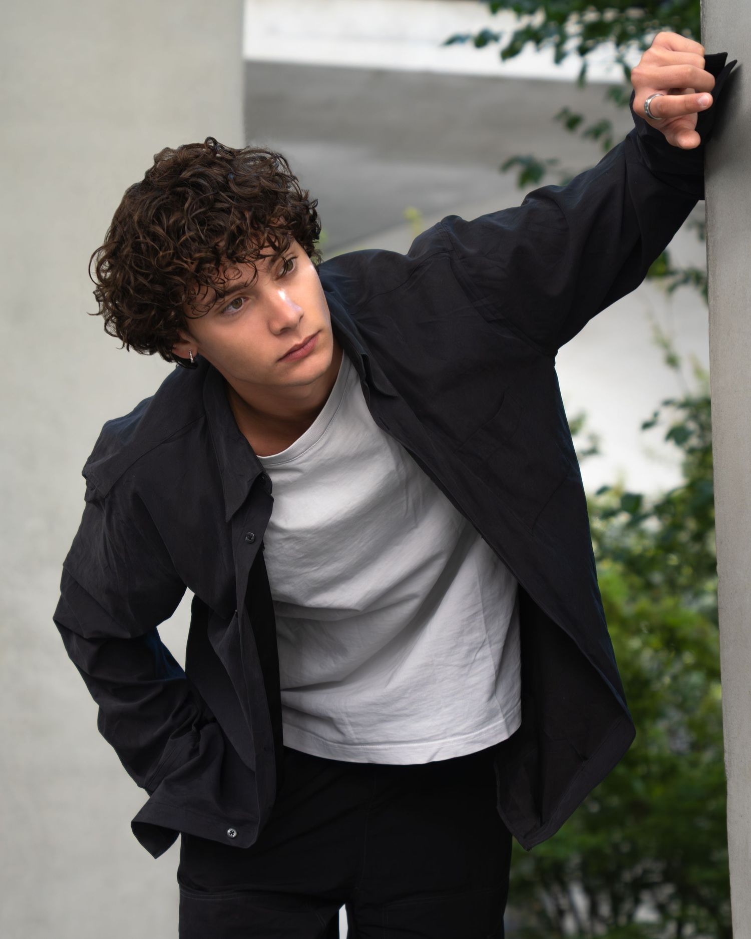 Someone in a black jacket and white t-shirt poses against a wall with greenery in the background.