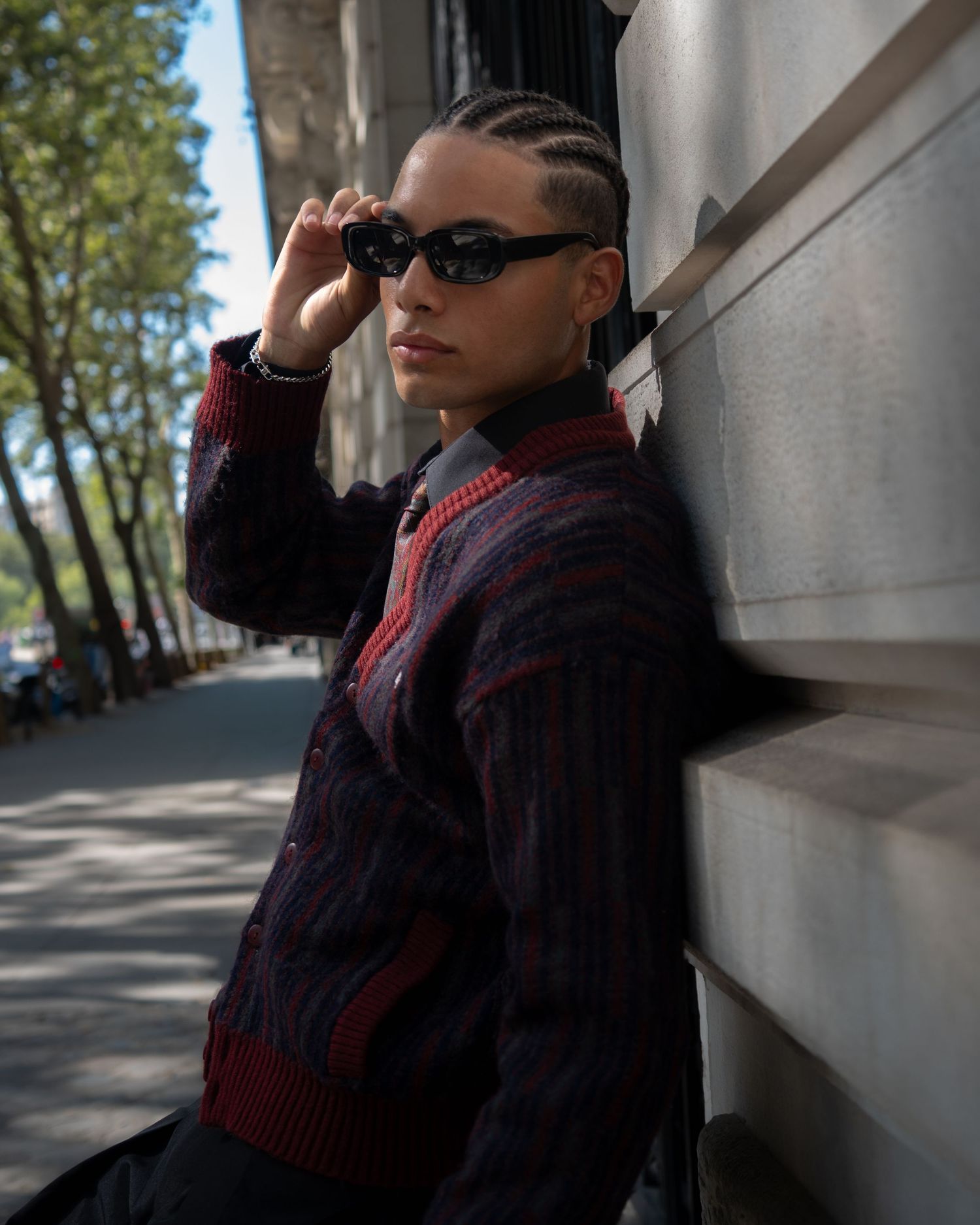 Person with cornrows wearing dark cardigan and black sunglasses leaning against wall on urban sidewalk.