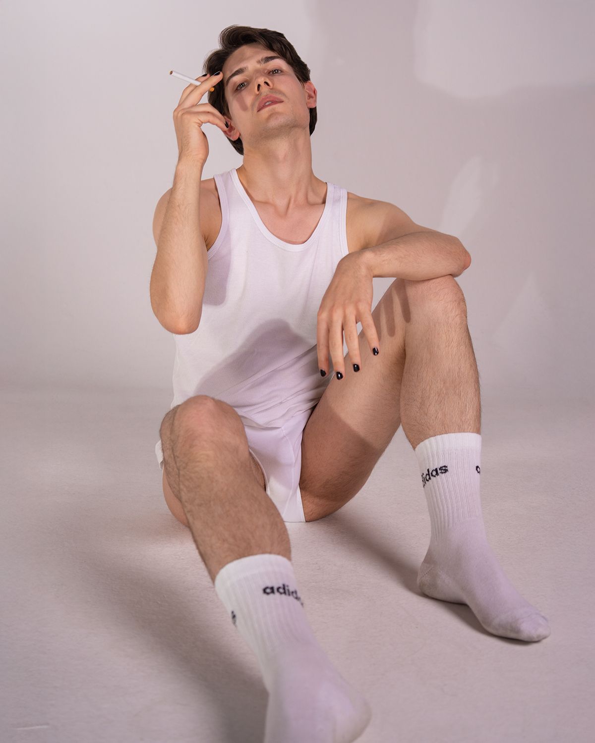 Person in white tank top and socks sits in artistic pose against light background during studio photoshoot.