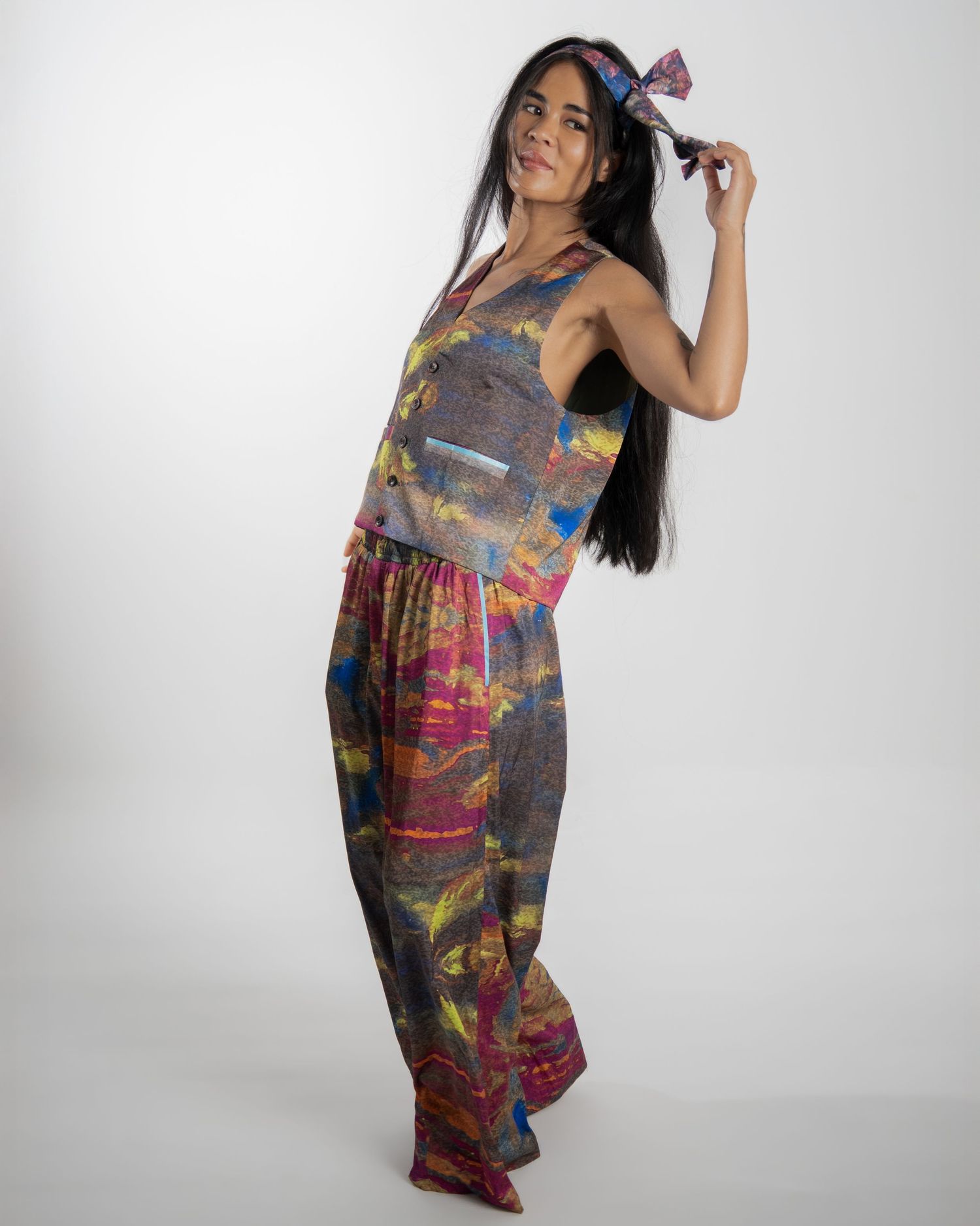 Woman wearing colorful tie-dye jumpsuit with psychedelic pattern poses against white background while playing with long hair.