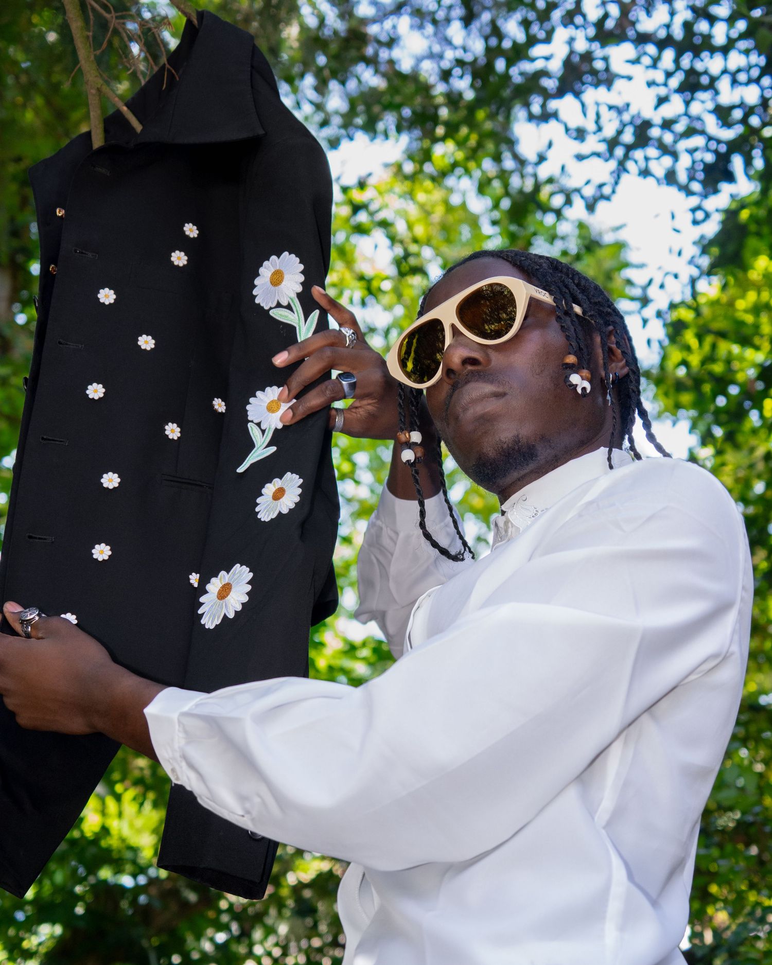 Person in white shirt and sunglasses holding up a black jacket with daisy print pattern against leafy outdoor background.