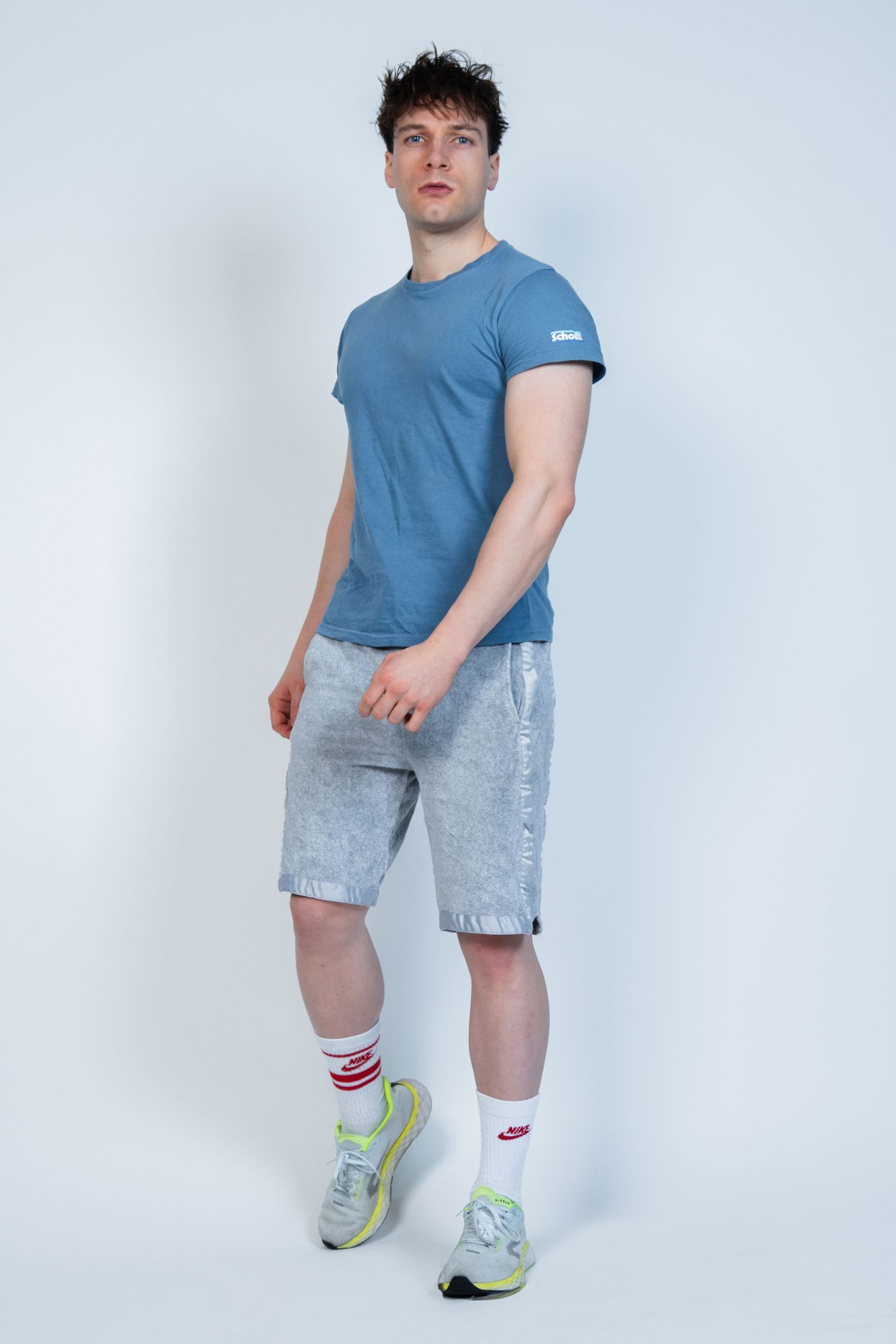 Modèle athlétique portant un t-shirt bleu, un short en jean délavé clair, des chaussettes rayées et des baskets vertes sur fond blanc.