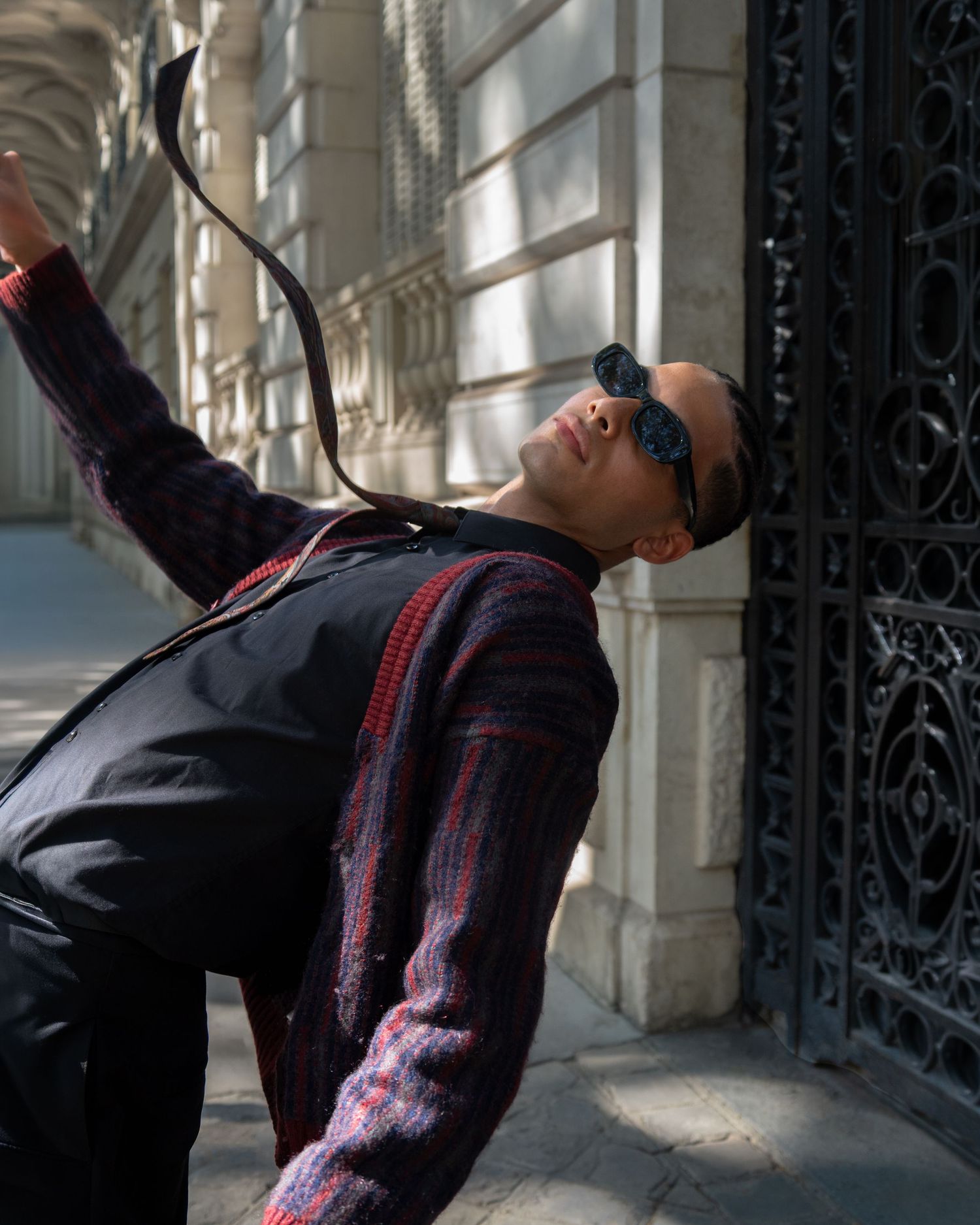 A stylish man in sunglasses and a dark patterned jacket poses dramatically against an ornate iron gate.