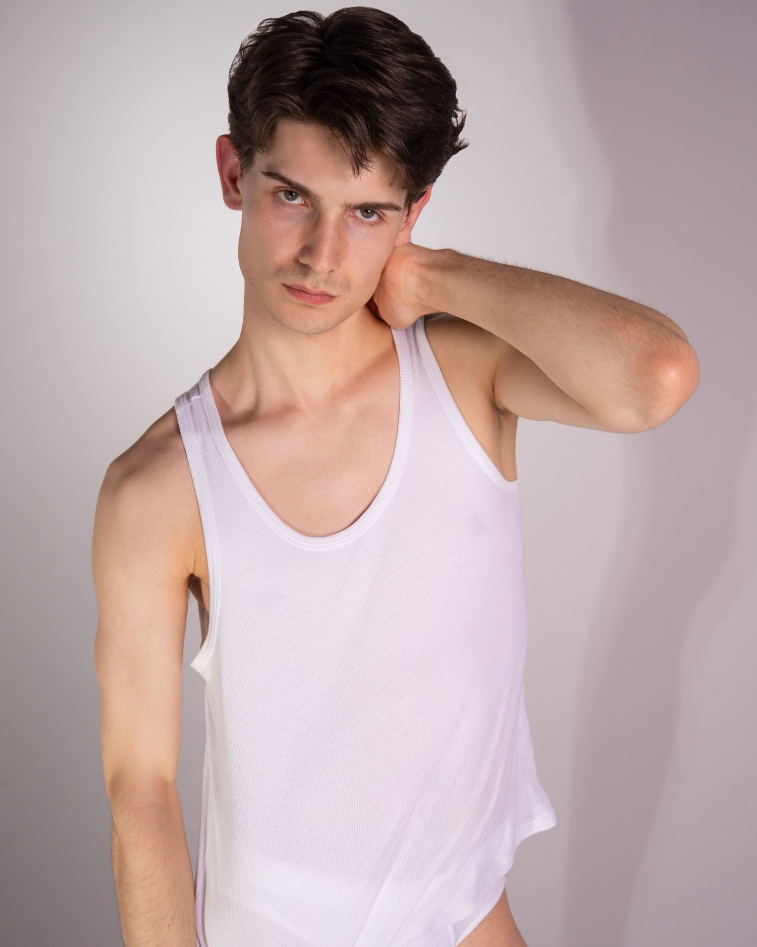 Person wearing a white sleeveless tank top poses with hand near neck against a light gray background.