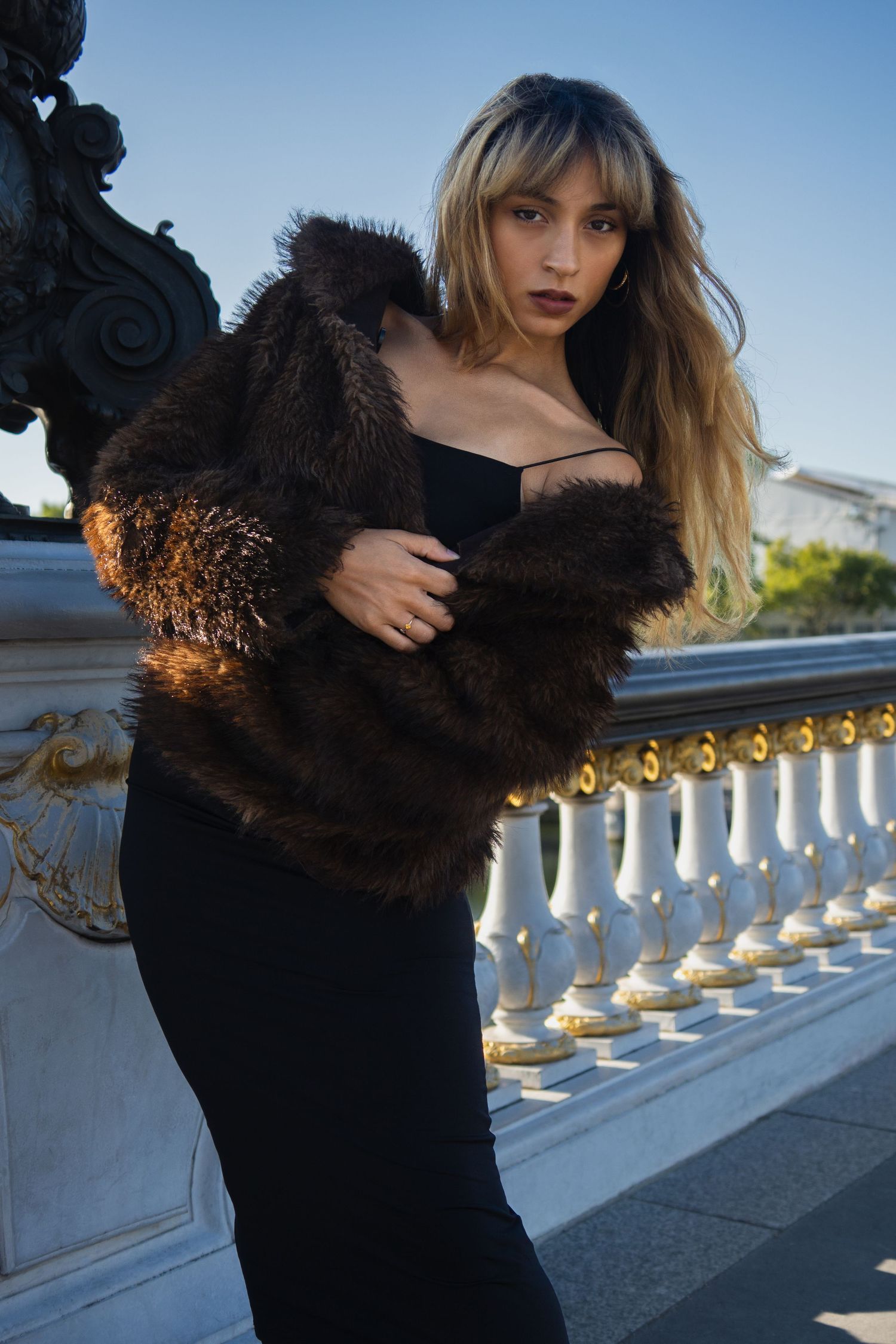 Woman in brown fur coat and black dress poses by ornate white balustrade under clear blue sky.