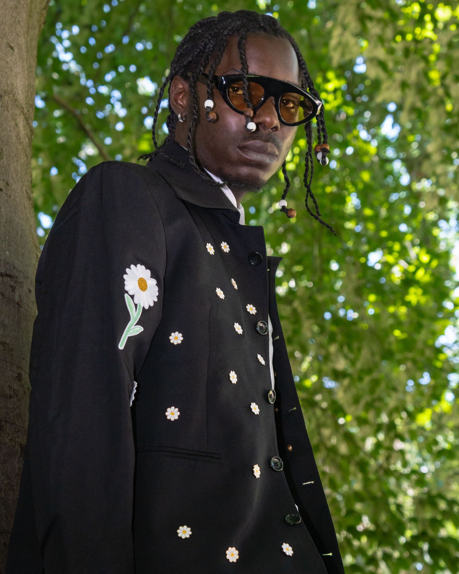 Fashion portrait showing a black coat adorned with daisy embroidery against a sunlit green backdrop.