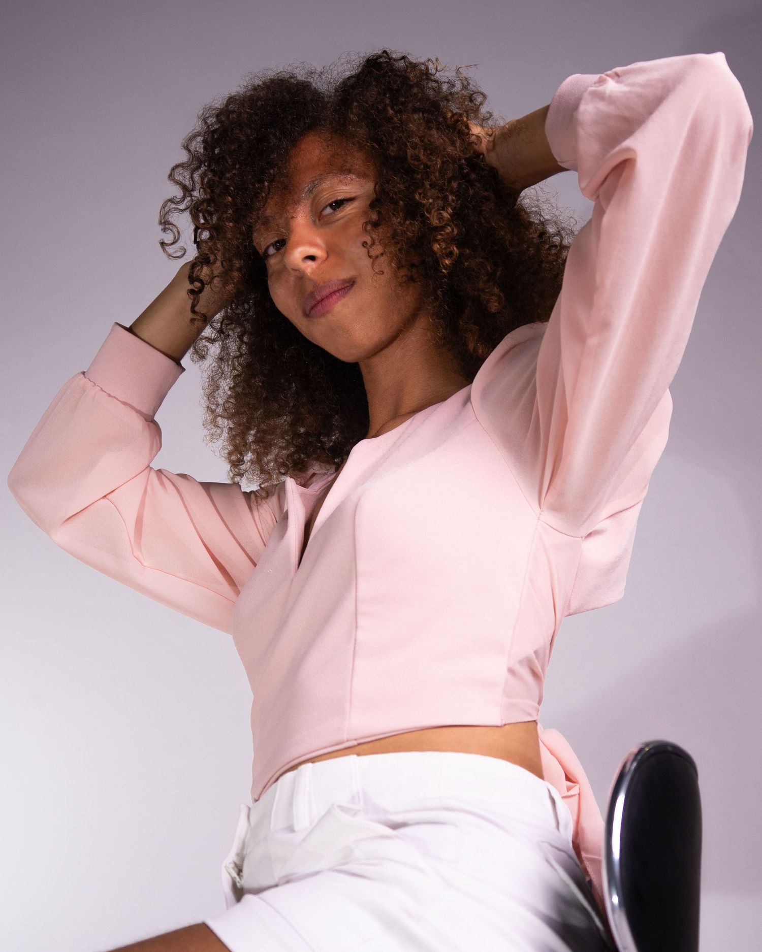 Person with curly hair wearing a pink cropped sweater poses gracefully against a light background.