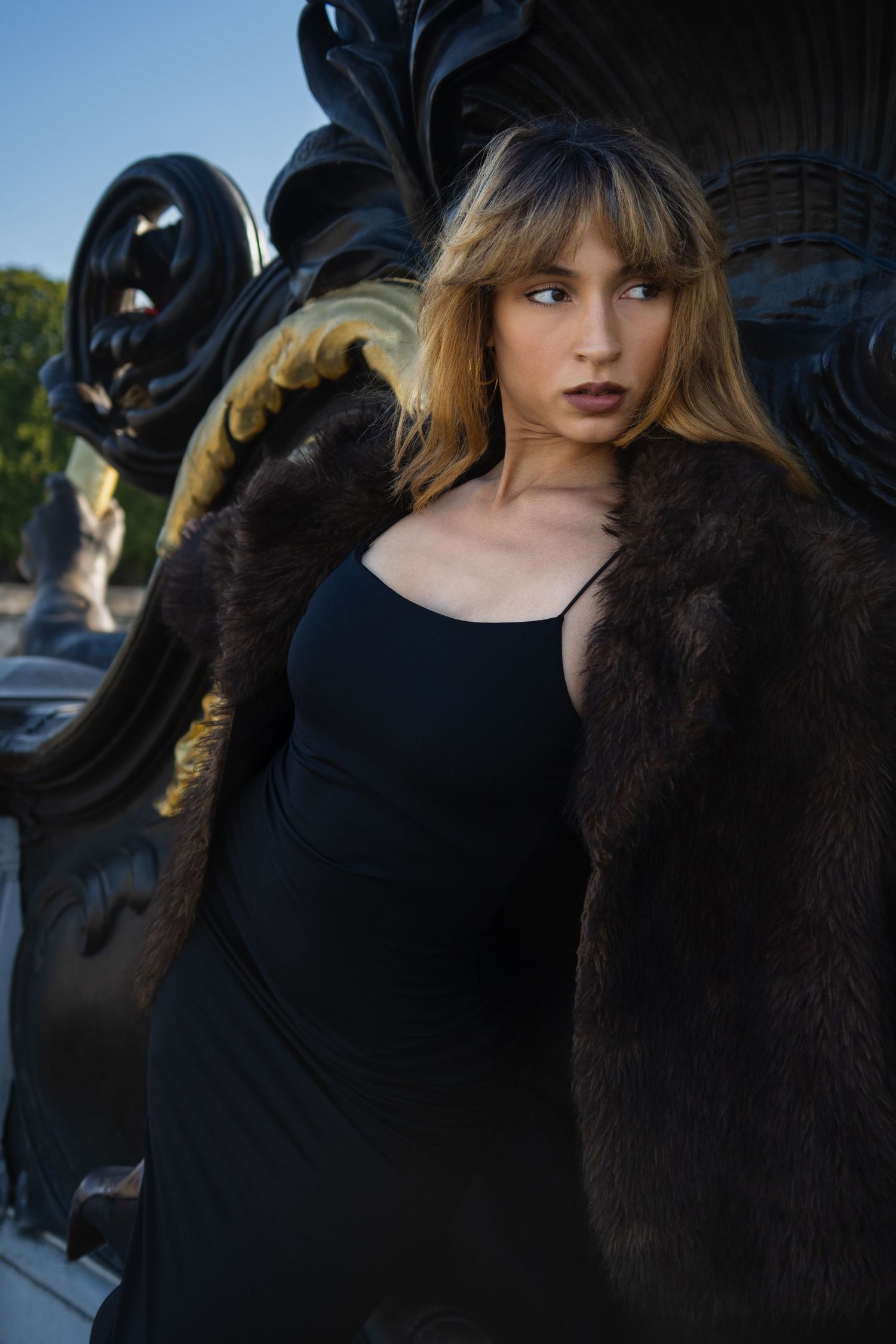 Young woman in black dress and brown fur coat poses elegantly against ornate dark sculpture outdoors.