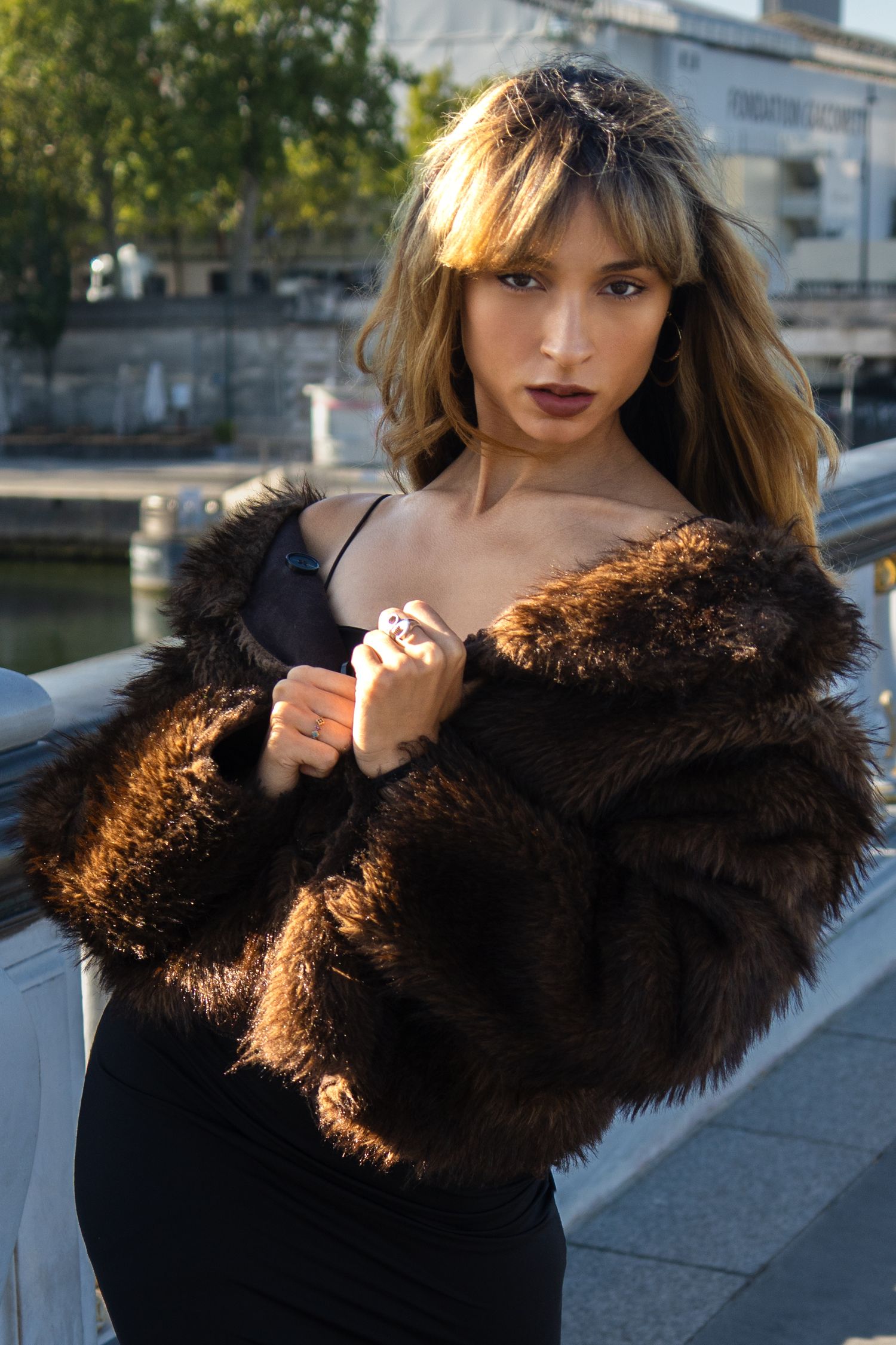 Woman in brown fur coat and black dress posing outdoors near a railing with urban background.