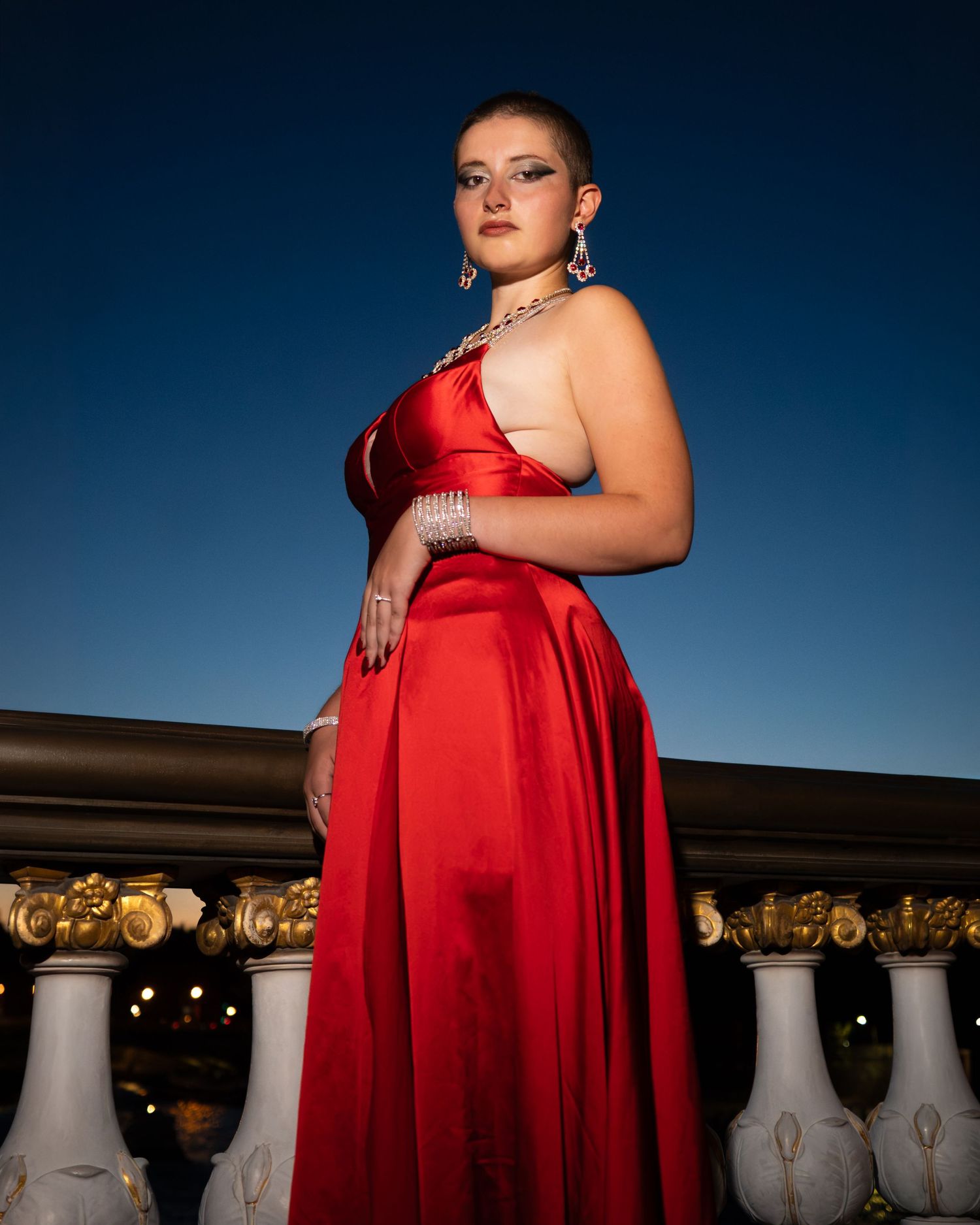Young woman in elegant red satin gown with silver jewelry poses confidently at dusk against ornate white balustrade.