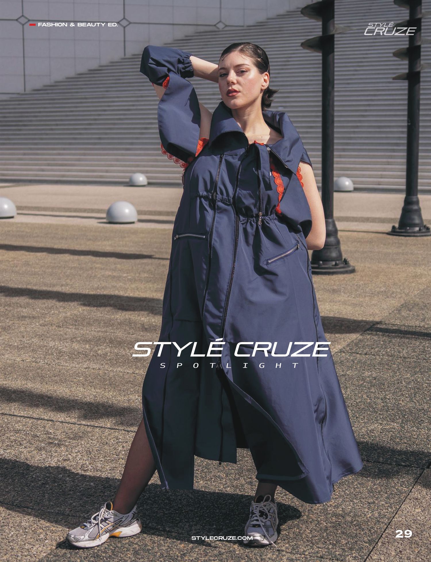 Model wearing navy blue oversized utility dress coat with red accents and sneakers on urban sidewalk with concrete spheres.