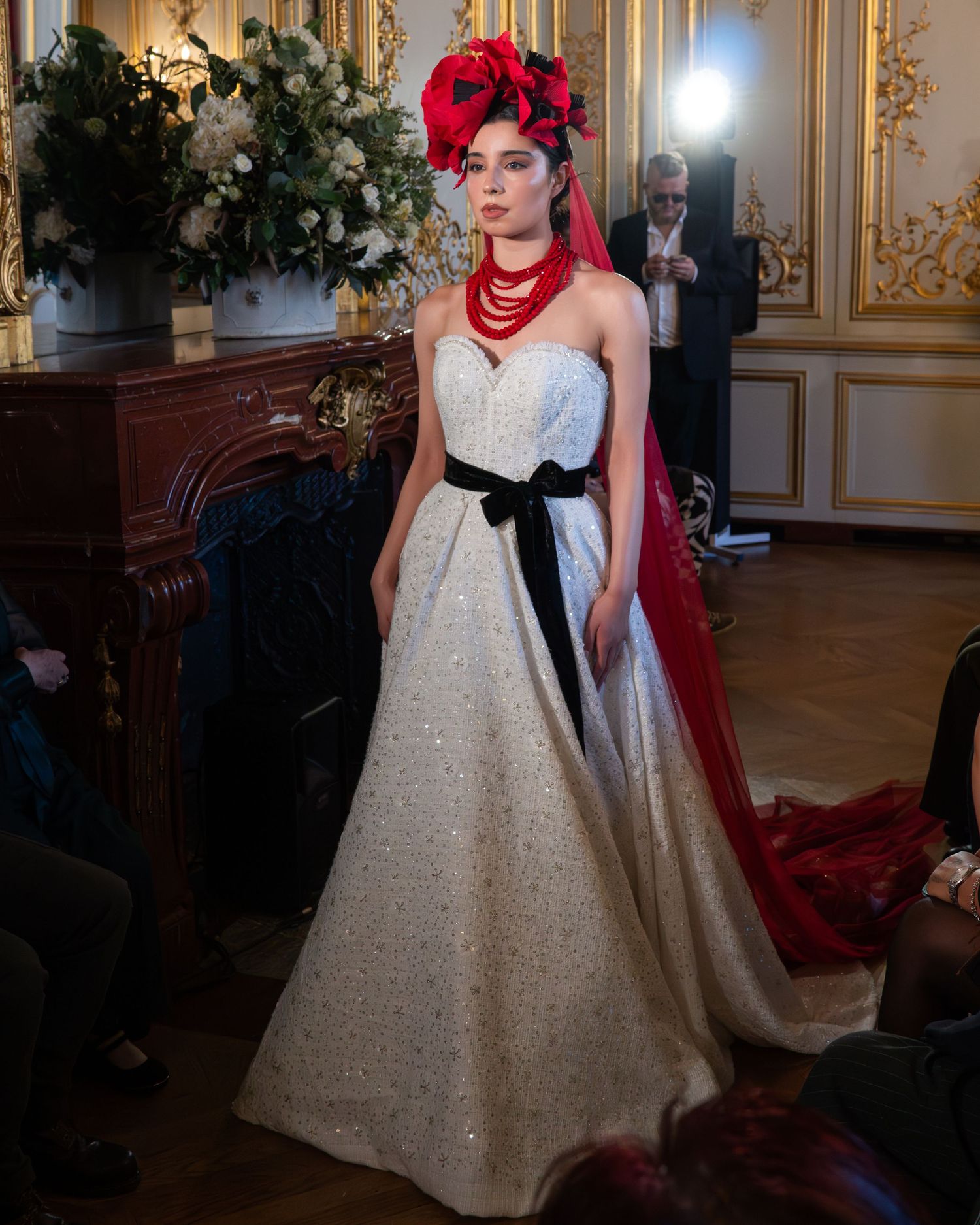 Mannequin portant une robe de bal blanche scintillante avec une ceinture noire et une coiffe rouge spectaculaire dans une pièce dorée ornée.