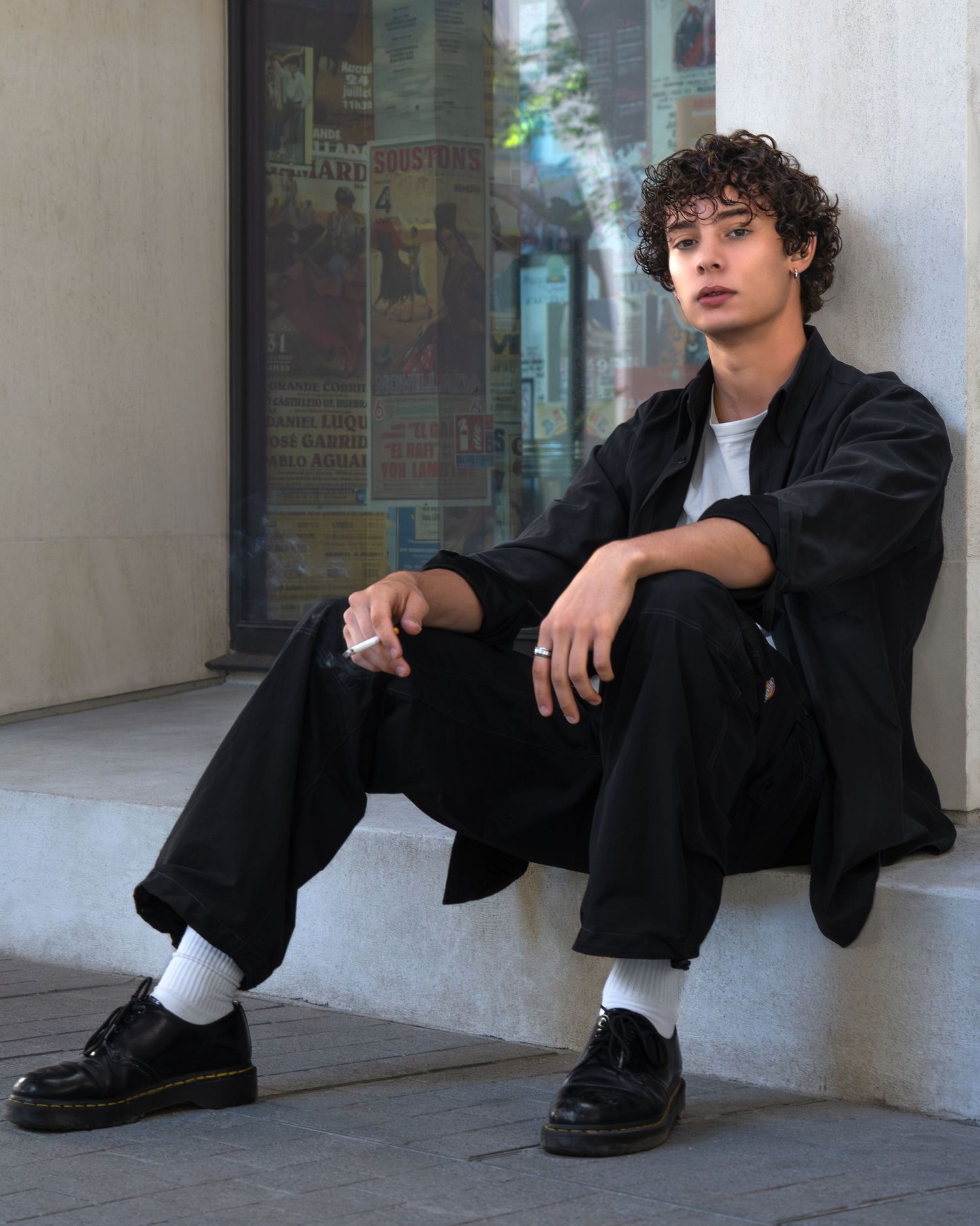 A figure in a black blazer and pants sits against a white wall while wearing shiny dress shoes and white socks.