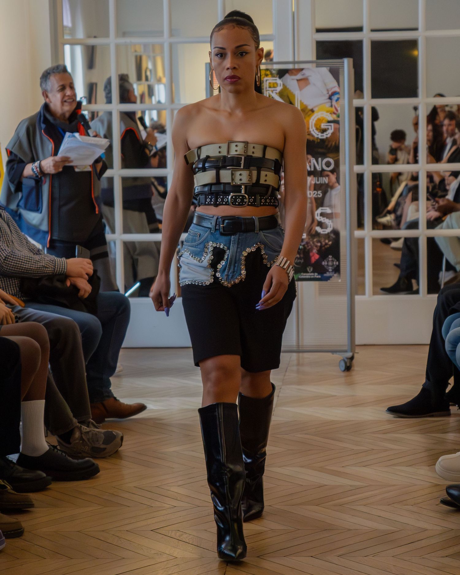 A fashion show model walks the runway wearing a denim and black ensemble with knee-high leather boots on a hardwood floor.