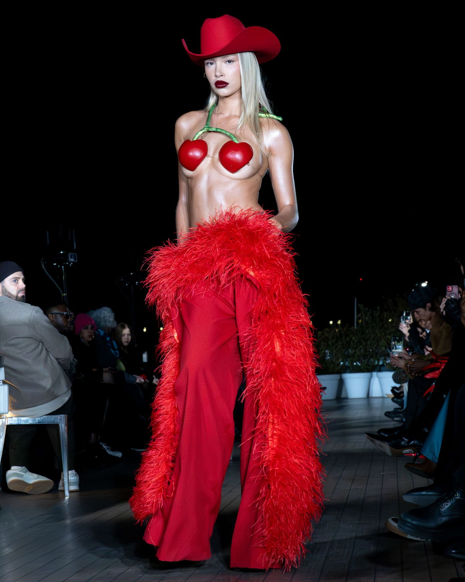 Mannequin en costume rouge avec des cache-cœurs et un boa à plumes sur le podium la nuit lors de la Paris Fashion Week pour AURELA HOXHA.