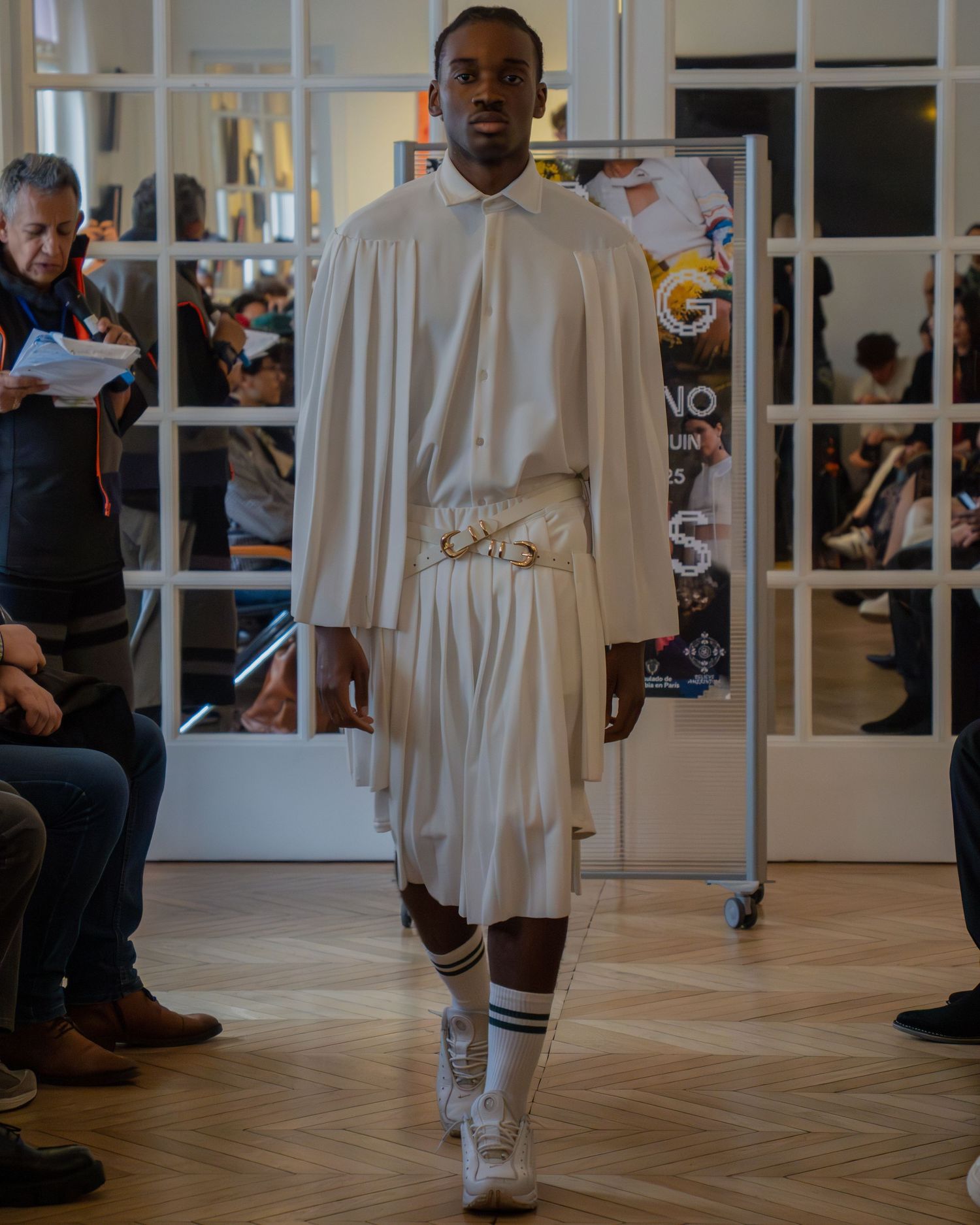Person wearing a flowing white robe walks across a polished floor in an elegant indoor space during a fashion event.