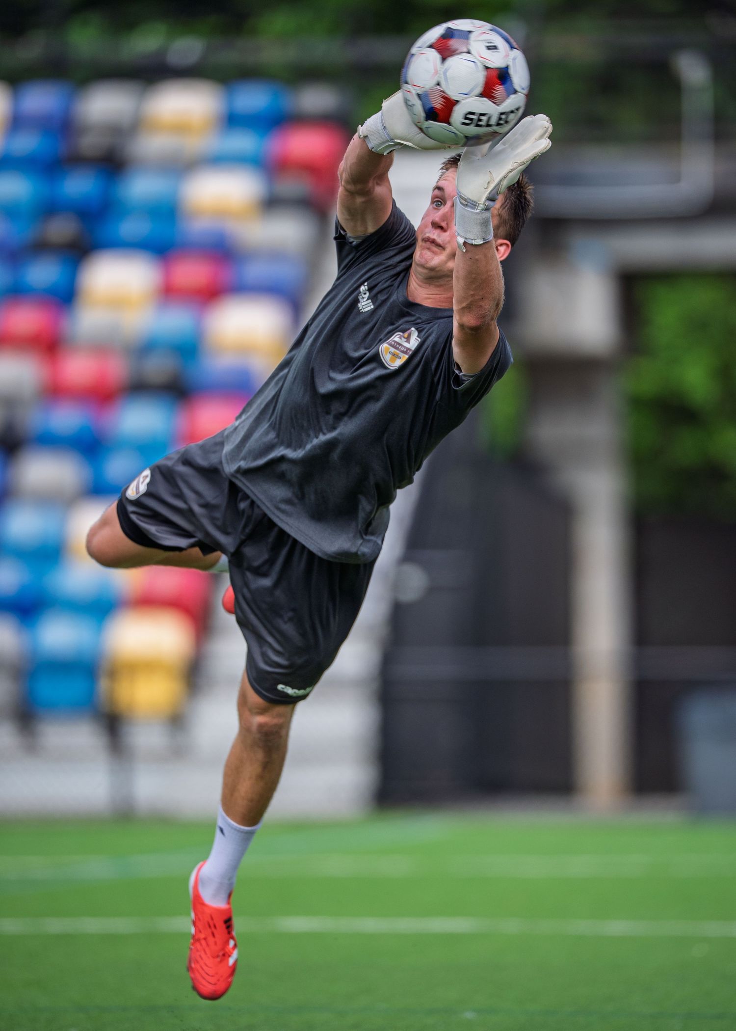 Soccer goalkeeper in black uniform leaps high to catch a ball during training at a colorful stadium setting.