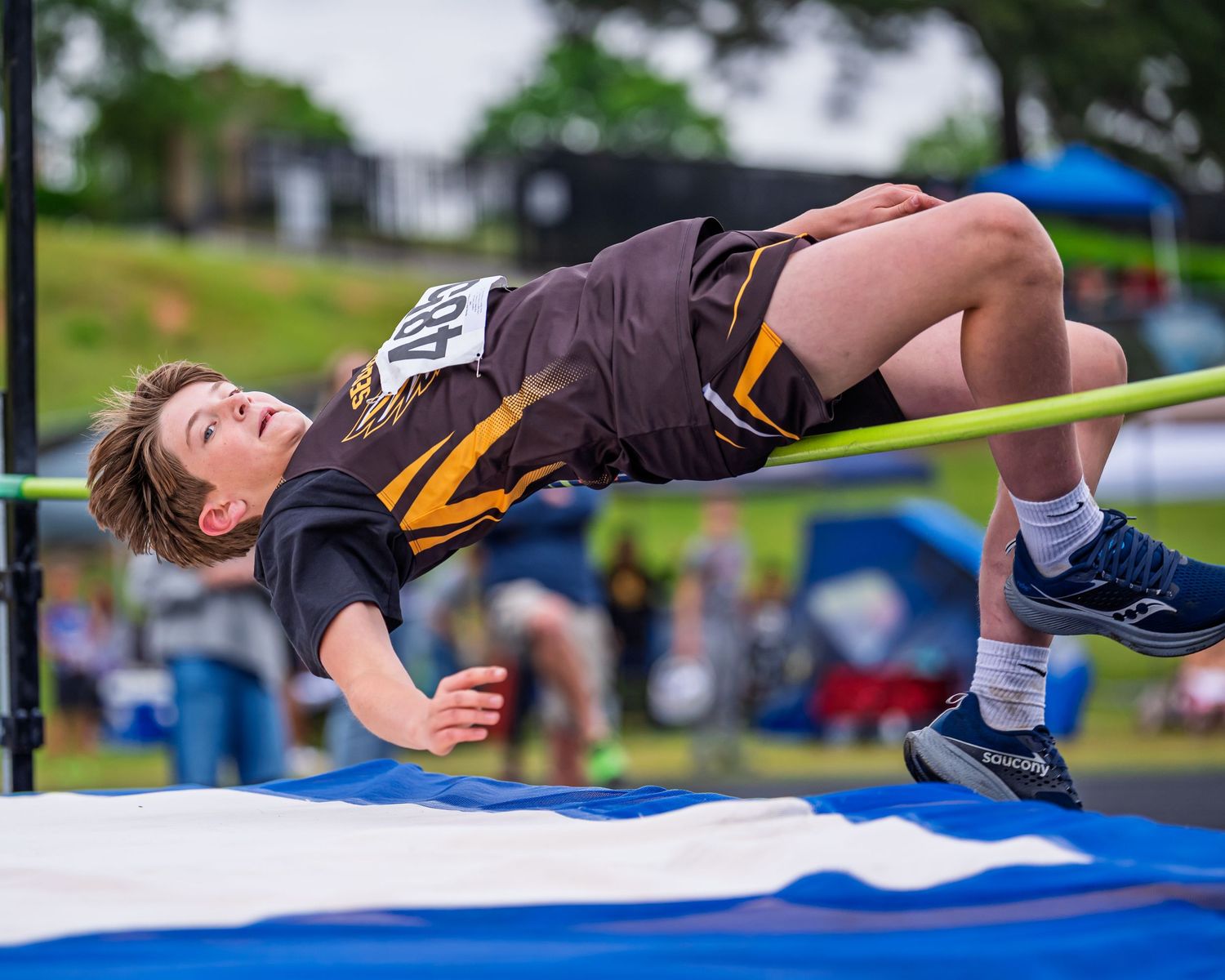 High school athlete performing high jump technique over crossbar at track and field competition with landing mat below.