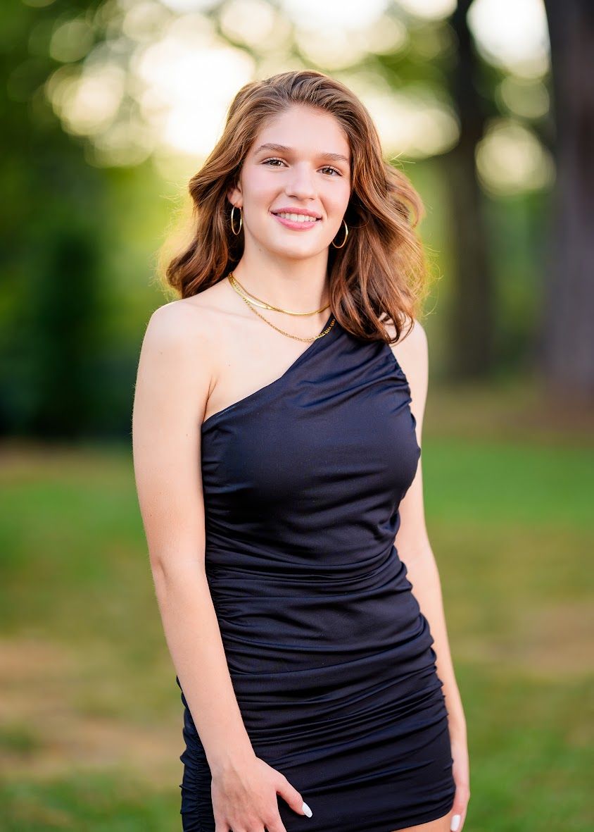 Portrait of a young woman in a black one-shoulder dress against a blurred green background.